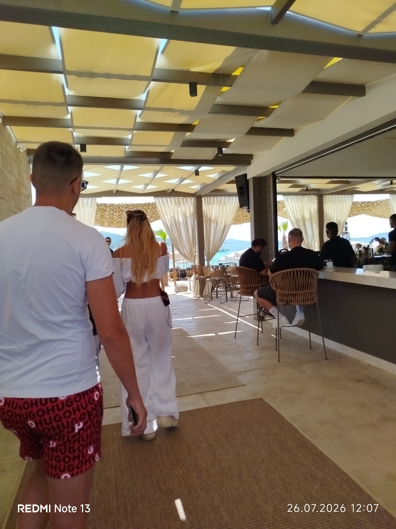 A man and a woman are walking away from the camera on a path at a beachside establishment in Sithonia Municipal Unit, Greece. The man, in the foreground, is wearing a white t-shirt and red shorts with a repeating "HUGO" pattern. The woman is wearing a white off-the-shoulder top and white wide-leg pants. In the background, a bar area is visible with several people seated on stools and standing behind the counter. Sunlight is filtering through a fabric canopy overhead, casting patterns of light and shadow on the floor. The environment appears to be a casual outdoor dining or lounge area, likely a beach club or restaurant, with a view of the sea and coastline in the distance.