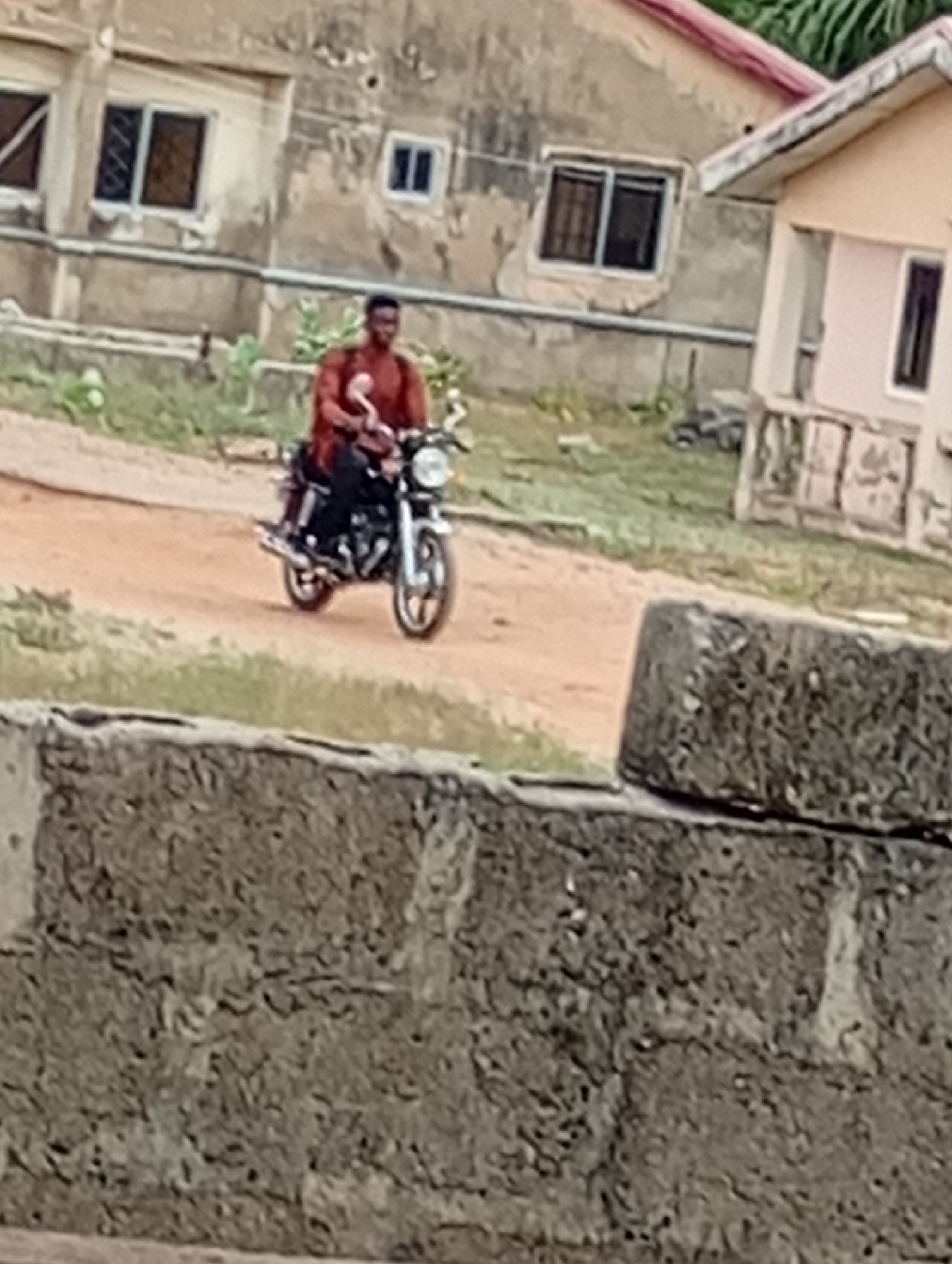 A man is riding a motorcycle on a dirt road. He is wearing a reddish-brown shirt and black pants. The motorcycle is black with silver accents. In the background, there are several buildings. A stone wall is in the foreground. Location is Unknown, Unknown.