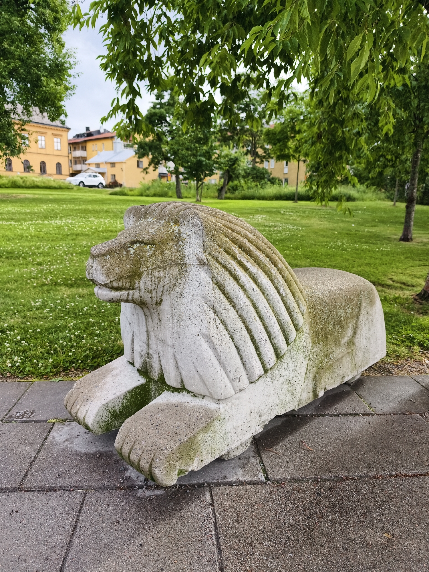 The image shows a stone lion statue in a park in Linköping, Sweden. The statue is weathered and covered in moss, suggesting it has been there for some time. The lion is depicted in a seated position, with its paws extended in front of it. The background shows a green lawn with scattered white flowers, trees, and buildings. The buildings appear to be residential or institutional, with yellow facades and red-tiled roofs. A white car is parked on the grass in the distance. The sky is overcast, indicating a cloudy day. The scene is peaceful and serene, suggesting a quiet moment in a public space.