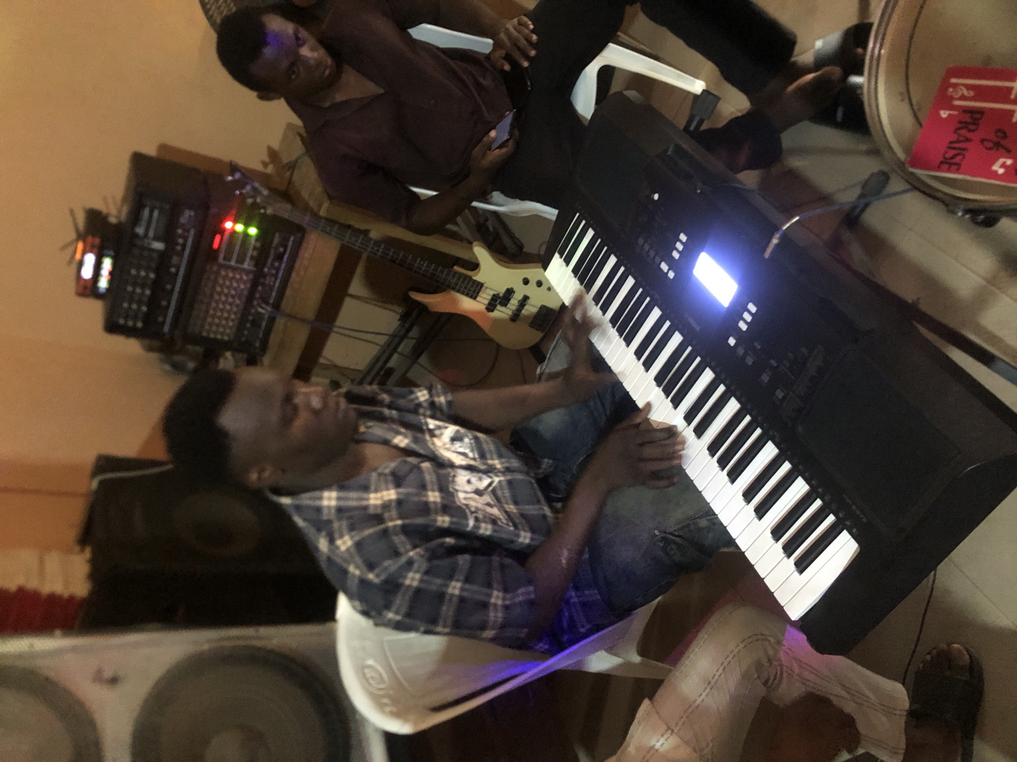 A slightly tilted, high-angle indoor shot features two men and a variety of musical equipment. One man, wearing a blue plaid shirt and jeans, sits on a white plastic chair and appears to be playing a large black keyboard. The second man, wearing a maroon collared shirt and dark pants, sits on another white plastic chair in the background, looking at a smartphone.
To the left of the keyboard, a bass guitar leans against a stack of electronic equipment. A drum set is visible to the right of the keyboard. A large speaker is visible at the bottom left corner. The location is Nkwelle Ezunaka, Nigeria, but the lack of exterior context prevents further specifics.
