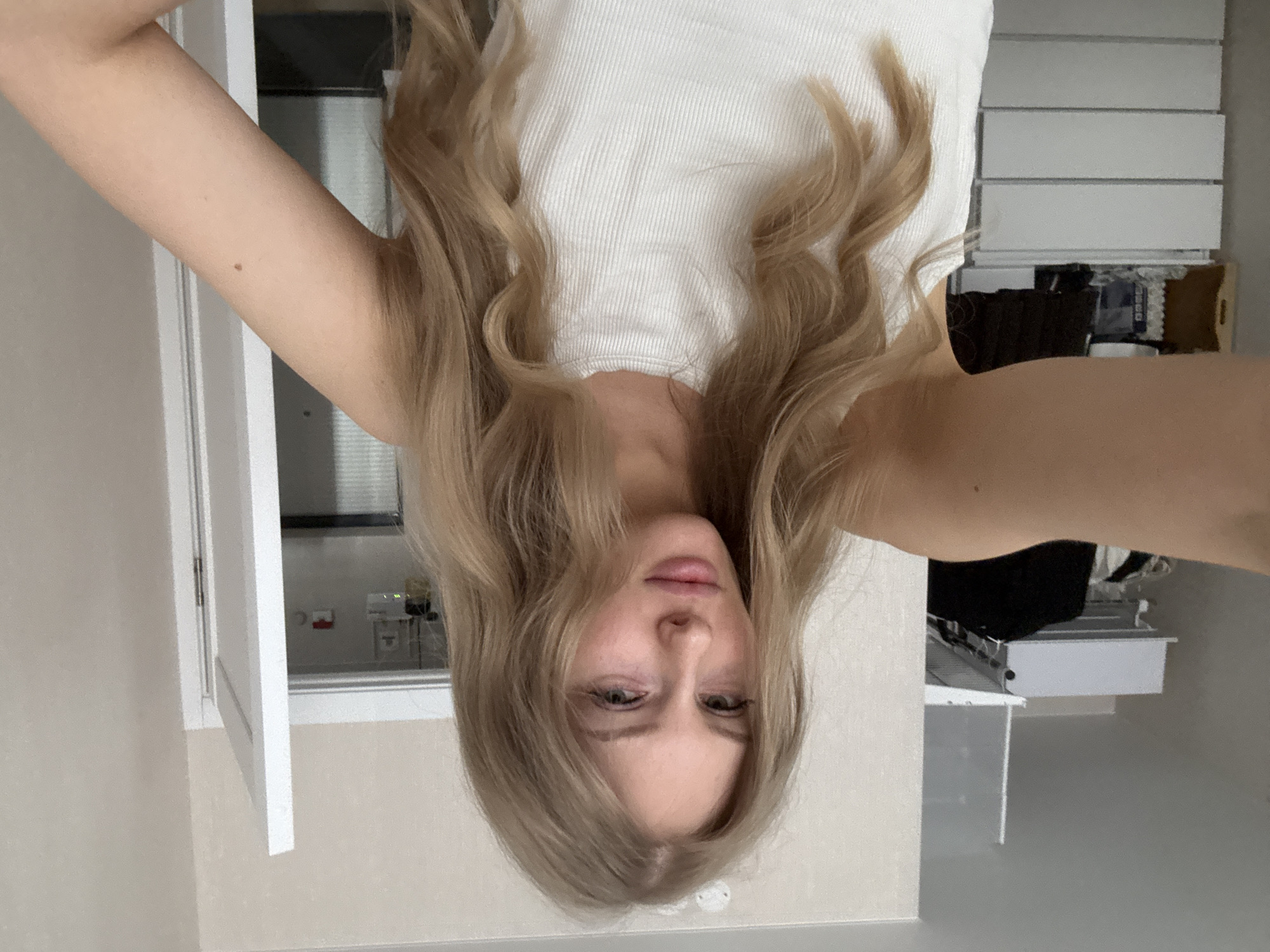 A young woman is holding the camera upside down. The subject appears to be inside a room with a white wall and white trim and a door. The subject is wearing a white ribbed tank top. The woman has long blonde wavy hair. A closet is visible behind the woman, with drawers and clothes inside. The location cannot be determined to be Perm, Russia based on the provided image.