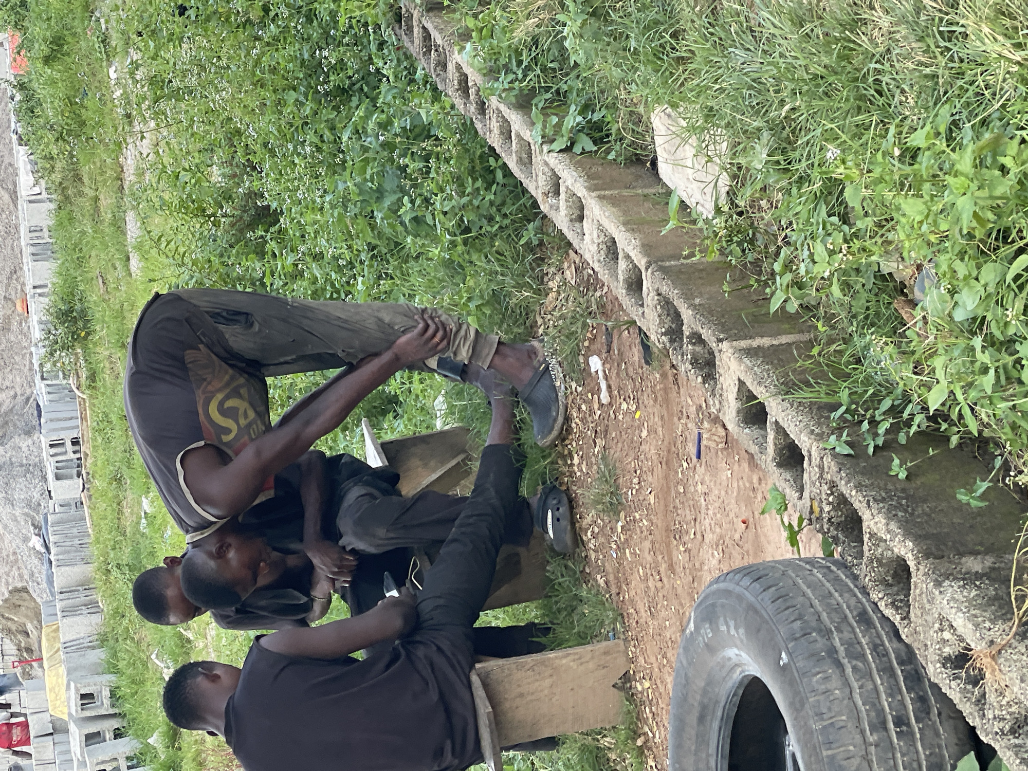 In an outdoor, unpaved area within Abuja, Nigeria, three young men are gathered. One man, wearing a dark t-shirt and pants, is bent over, attending to his foot or shoe, which is a dark-colored clog. He has a gold-colored chain visible around his neck, and his pants have a cargo pocket with a yellow and black design. He is leaning on a wooden bench where two other men are seated. The man on the left, wearing a black sleeveless shirt and black pants, faces away from the camera. The man in the middle, in a black t-shirt and black pants, faces towards the man tending to his foot. Behind the men, stacks of gray concrete blocks and mounds of dirt are visible, suggesting a construction site or material storage area. The ground is covered with reddish-brown soil, scattered with small stones and dried leaves. To the right, a low concrete structure, possibly a drainage channel or border, is lined with green vegetation and tall grass. In the foreground on the left, a deflated black tire is partially visible, resting on the ground. The overall scene depicts a moment of rest or informal gathering amidst a seemingly utilitarian or developing environment.
