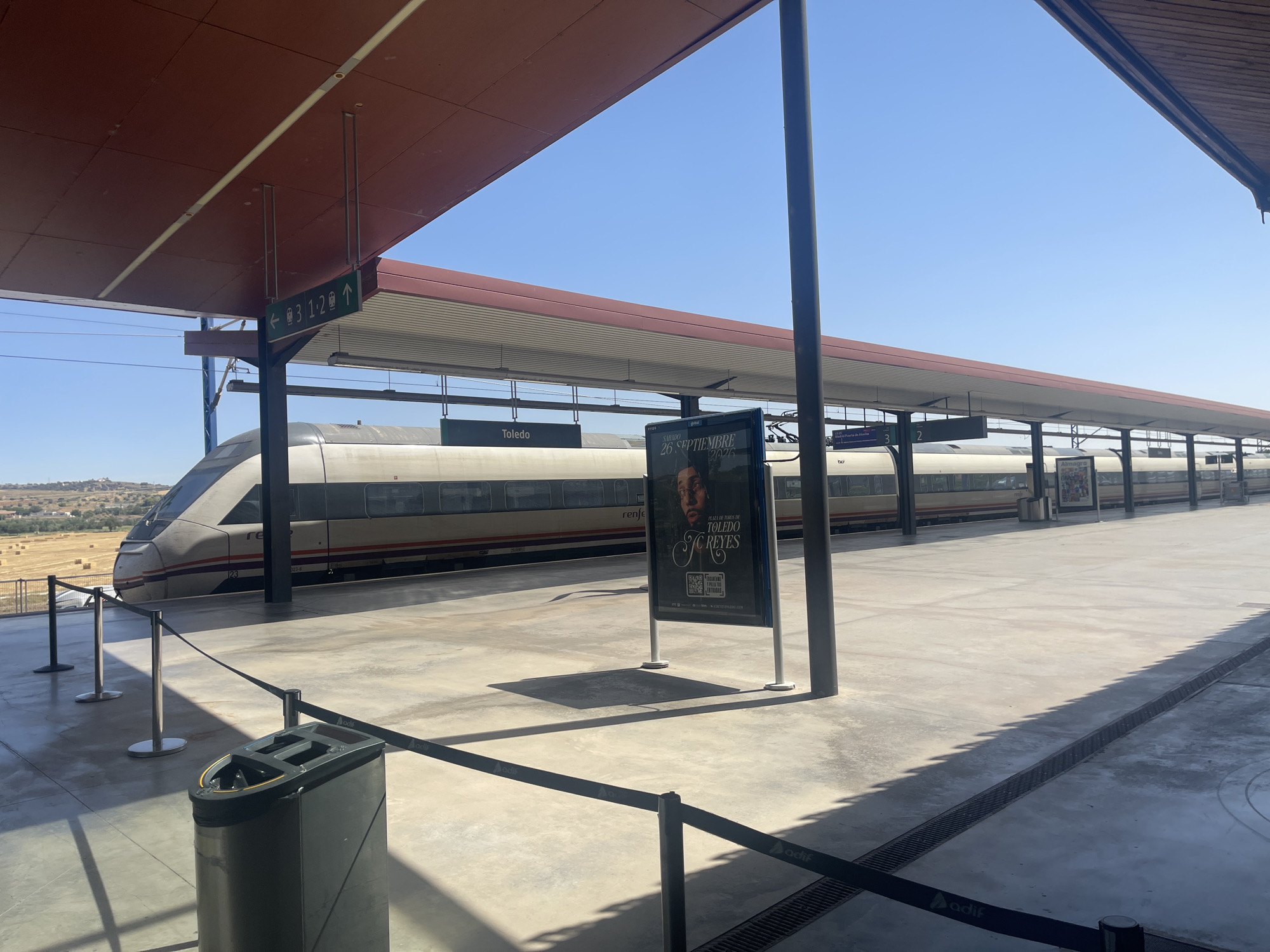 The image depicts a view of a train station platform in Toledo, Spain, on a clear, sunny day. A modern, high-speed train with the "renfe" logo is parked at the platform. The platform is covered by a red canopy, and there are signs indicating track numbers and directions. One sign points left with an arrow and a train icon, indicating track 3, and another points right with an arrow and train icons, indicating tracks 1 and 2. A large advertising billboard is also present on the platform. In the background, beyond the train, there are fields and hills under a bright blue sky. The platform itself is paved with concrete and has a drainage grate running along its edge. Safety barriers are set up near the edge of the platform. A trash receptacle is visible in the foreground.