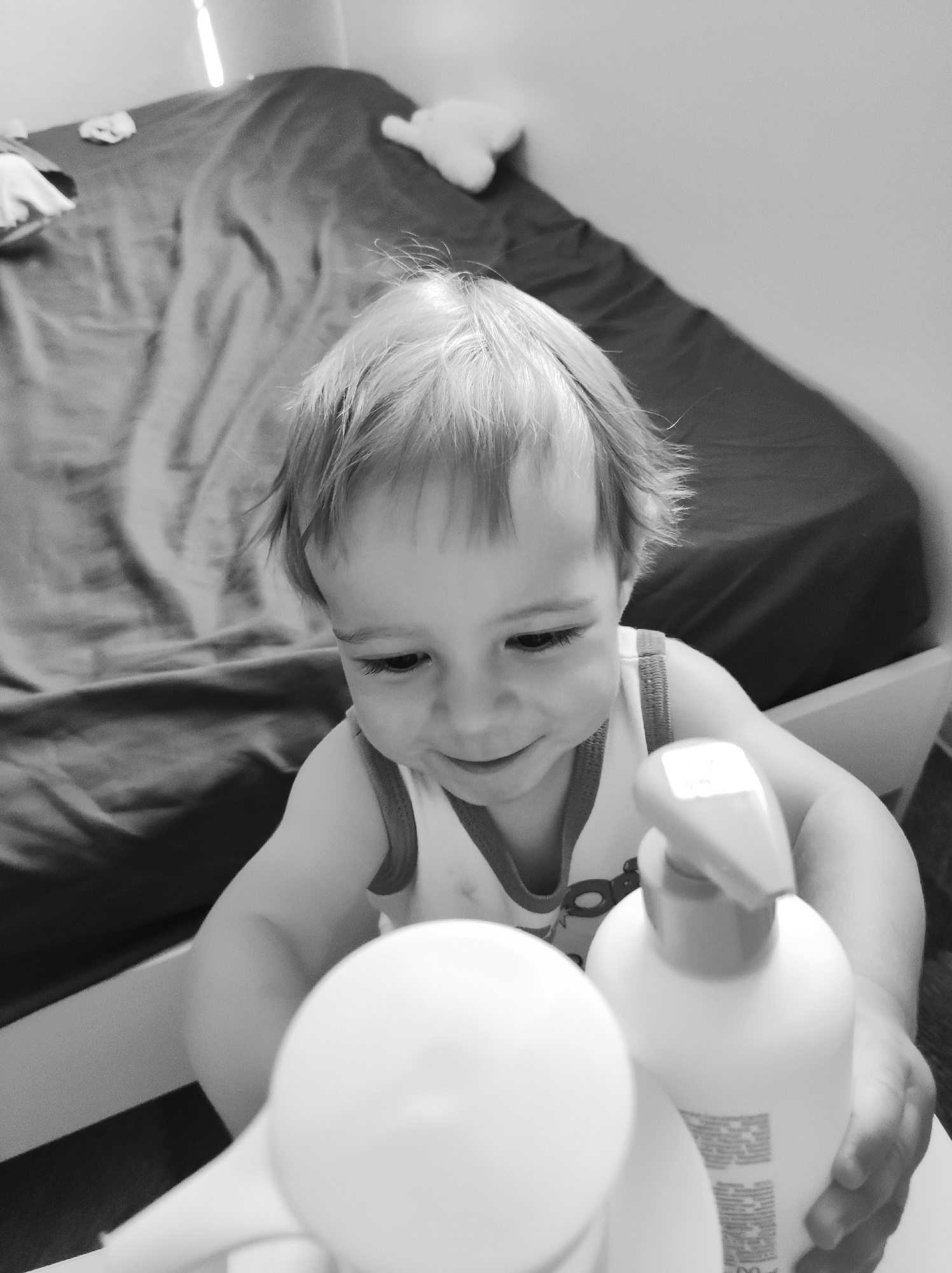A black and white close-up shot captures a toddler seated at a table, likely indoors near a bed. The toddler has light-colored hair and is wearing a tank top. They are interacting with a white pump-style bottle and a partially visible round object, possibly another bottle. The background features a bed with a dark-colored, rumpled cover. There are faint outlines of what appears to be a plush toy and other items on the bed. The location is specified as the city of Chaikovsky, Russia, although this cannot be verified from the visual information alone.