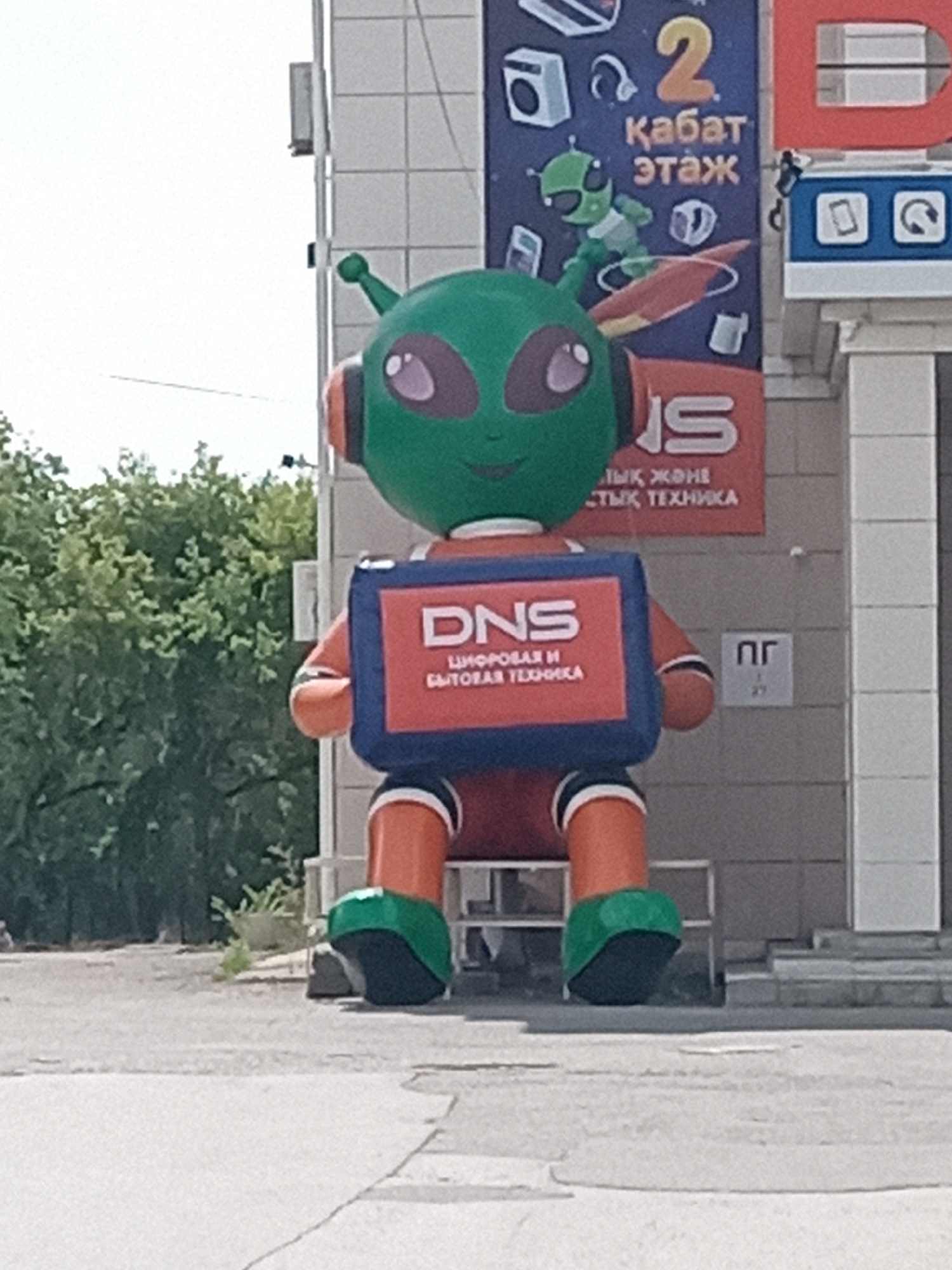 The image shows a large, inflatable alien mascot outside a building in Rudny, Kazakhstan. The mascot is green with large purple eyes and is wearing headphones. It is holding a blue and red sign that says "DNS" in white letters and "ЦИФРОВАЯ И БЫТОВАЯ ТЕХНИКА" below. Behind the mascot, a building has a large red "D" and part of an "N" visible. A sign with a cartoon alien riding a rocket and the number "2" with "Кабат этаж" (which translates to "Floor") is also visible. The environment appears to be an outdoor, urban setting with trees and pavement.