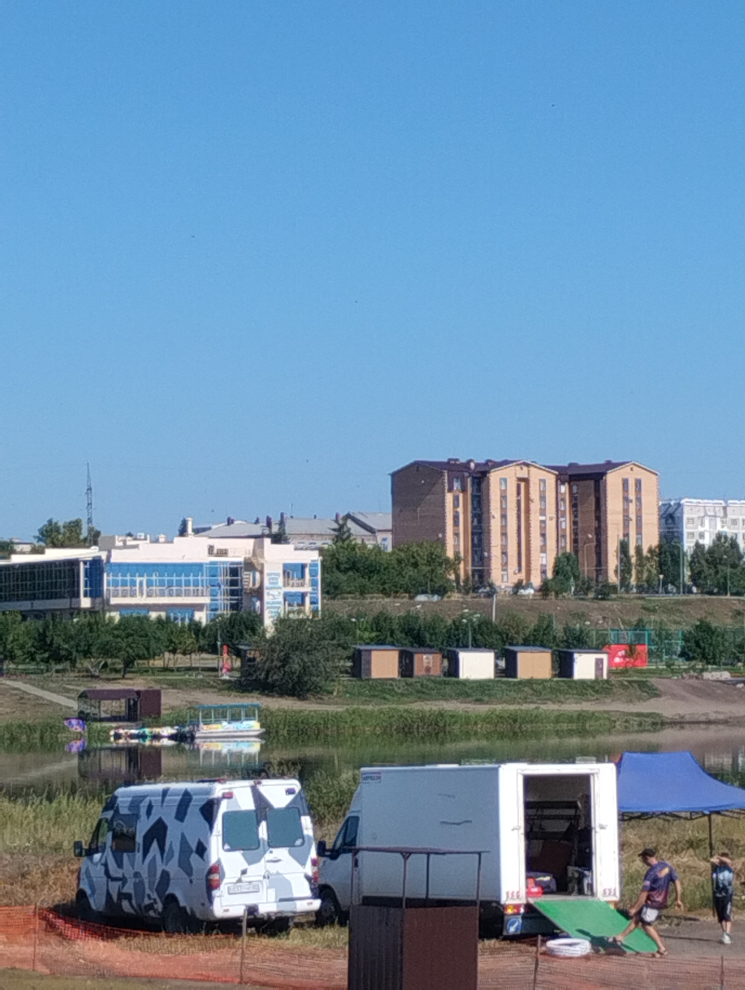 The image captures a waterfront area in Kostanay, Kazakhstan, on a clear day with a bright blue sky. In the foreground, an orange construction net is visible. To the left, a white van with a camouflage pattern is parked on a grassy embankment. Further to the right, another white van is partially visible, with its rear doors open, revealing equipment and a ramp. Two individuals are near this van; one is walking towards it, and another is standing to the right, looking away. A blue canopy tent is set up behind the vans.
The middle ground features a body of water, likely a river or lake, with two boats moored near a small, covered dock on the left. On the bank opposite the boats, a row of small, shed-like structures are lined up. In the background, there are several buildings. On the left, a modern building with large glass windows and blue accents stands out. To the right of this, a tall, multi-story apartment building with a brown roof and yellow-toned facade is prominent, with other residential buildings visible behind it. The overall scene suggests an outdoor recreational or event setup near urban development.