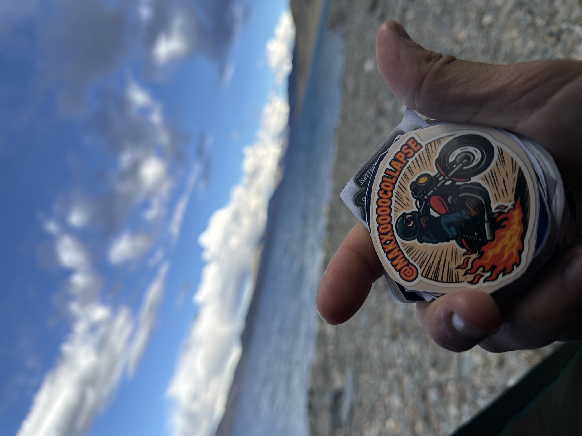 A person's hand is holding a stack of stickers against a blurred background of a body of water and mountains under a cloudy sky. The topmost sticker is round and features a cartoon illustration of a person riding a motorcycle with flames erupting from beneath it. The sticker also has text that reads "@MAXXOOOCOLLAPSE". The environment appears to be outdoors in a natural setting, possibly near a lake or a large body of water in a mountainous region. The lighting suggests it is daytime.