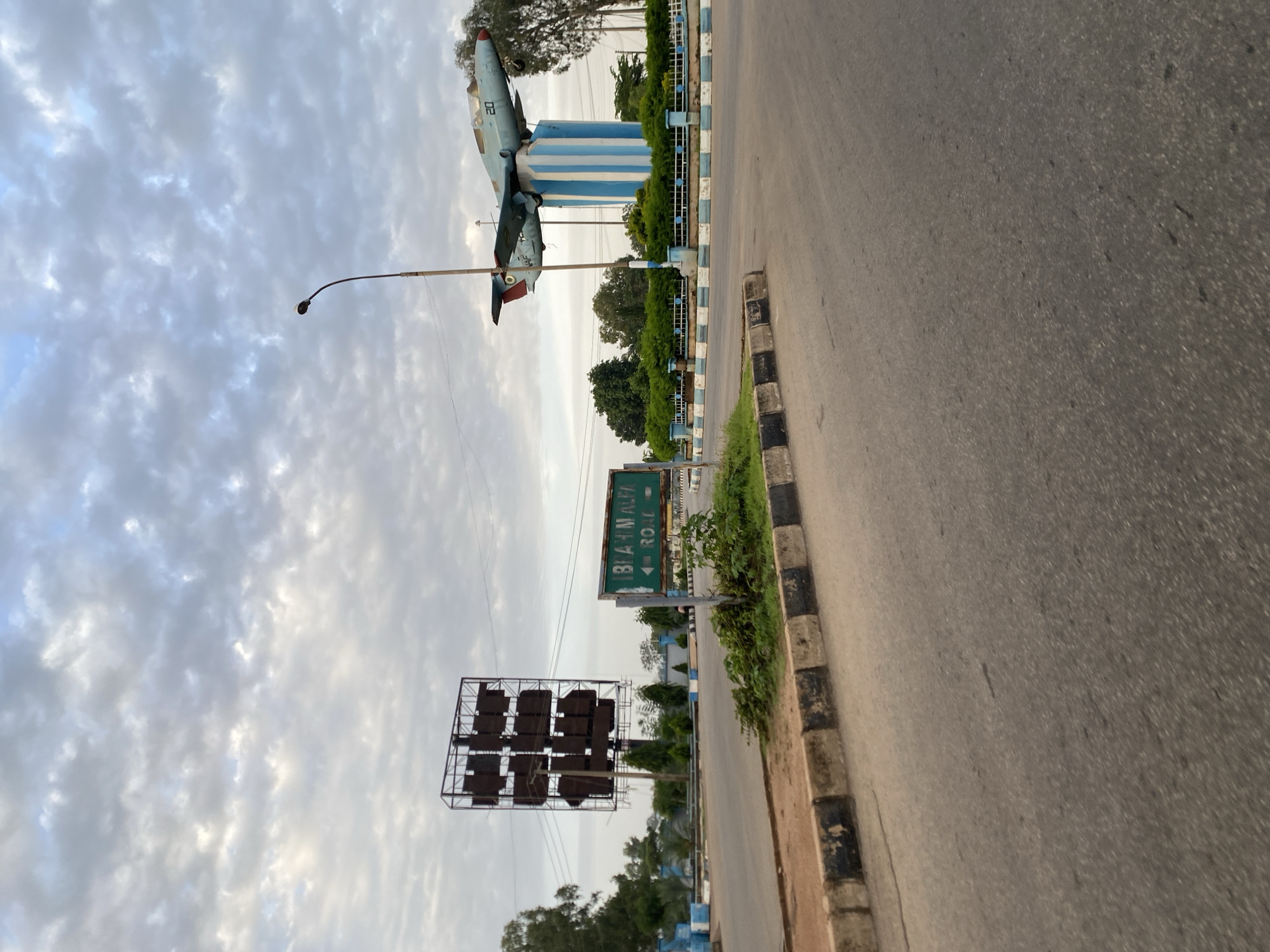 The image depicts a roundabout in Barakin Lerawi, Nigeria, featuring an airplane monument. The sky is overcast with textured clouds. On the right side of the roundabout, a light blue jet airplane, marked with the number "20" and some other insignia, is mounted on a cylindrical pedestal painted with alternating blue and white stripes. The airplane monument is surrounded by a low fence and dense green bushes. To the left of the airplane monument and closer to the viewer is a green road sign pointing left with the text "IBRAHIM ALFA ROAD". In the background, to the left of the sign, is a large, dark, rectangular billboard structure made of metal poles, with multiple square sections, appearing to be old or disused. The road itself is asphalt with clear lane markings. The roundabout is bordered by a curb with black and white painted sections. Trees are visible in the background on both sides of the road.