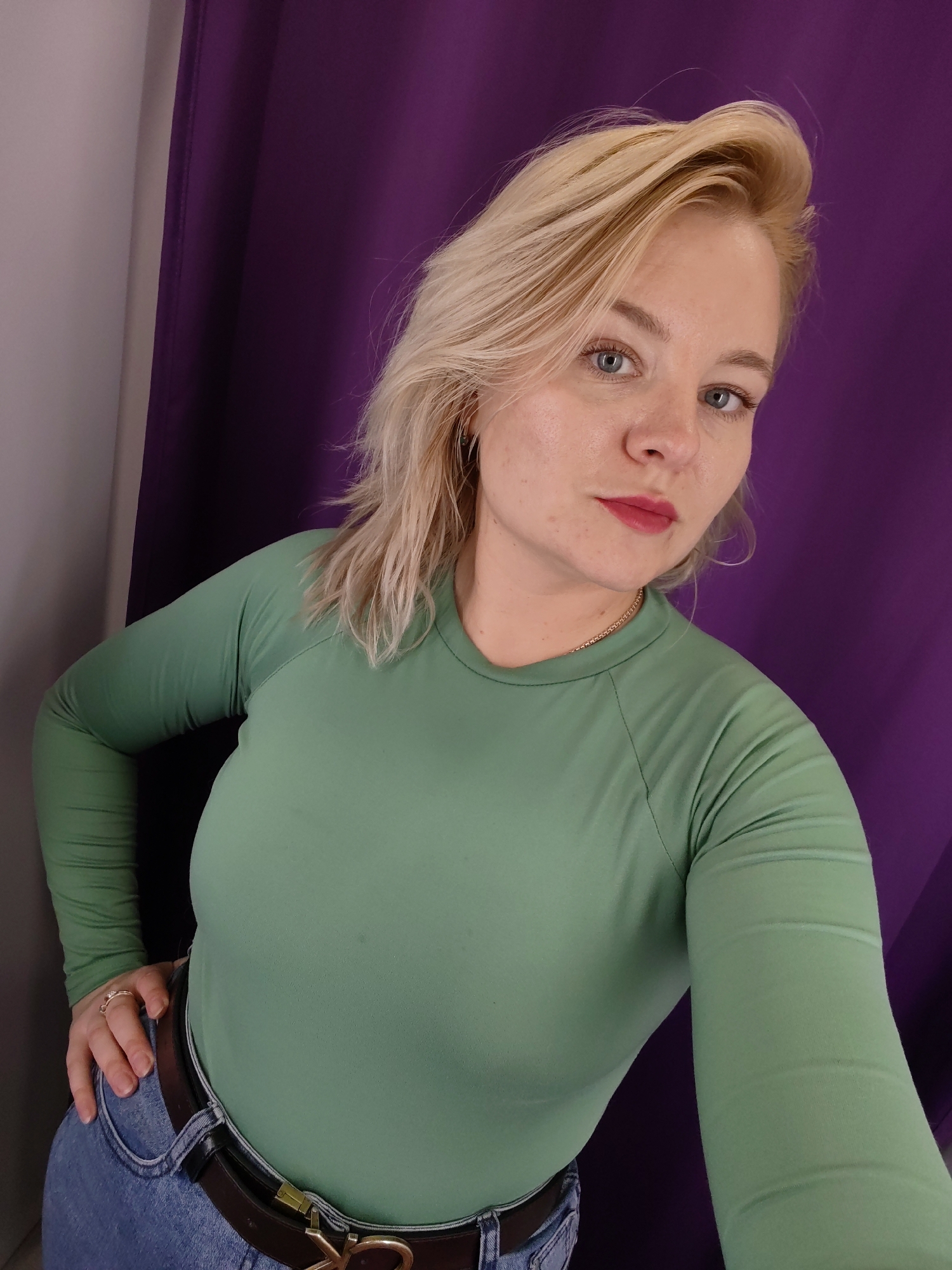 A fair-skinned woman with blonde hair and blue eyes is positioned against a backdrop of a purple curtain and a light gray wall. She is wearing a long-sleeved, light green top, blue jeans, and a dark brown leather belt with a gold-colored buckle. She has a gold chain around her neck and a ring on her left ring finger. Her left hand is resting on her hip. The location is Izhevsk, Russia.