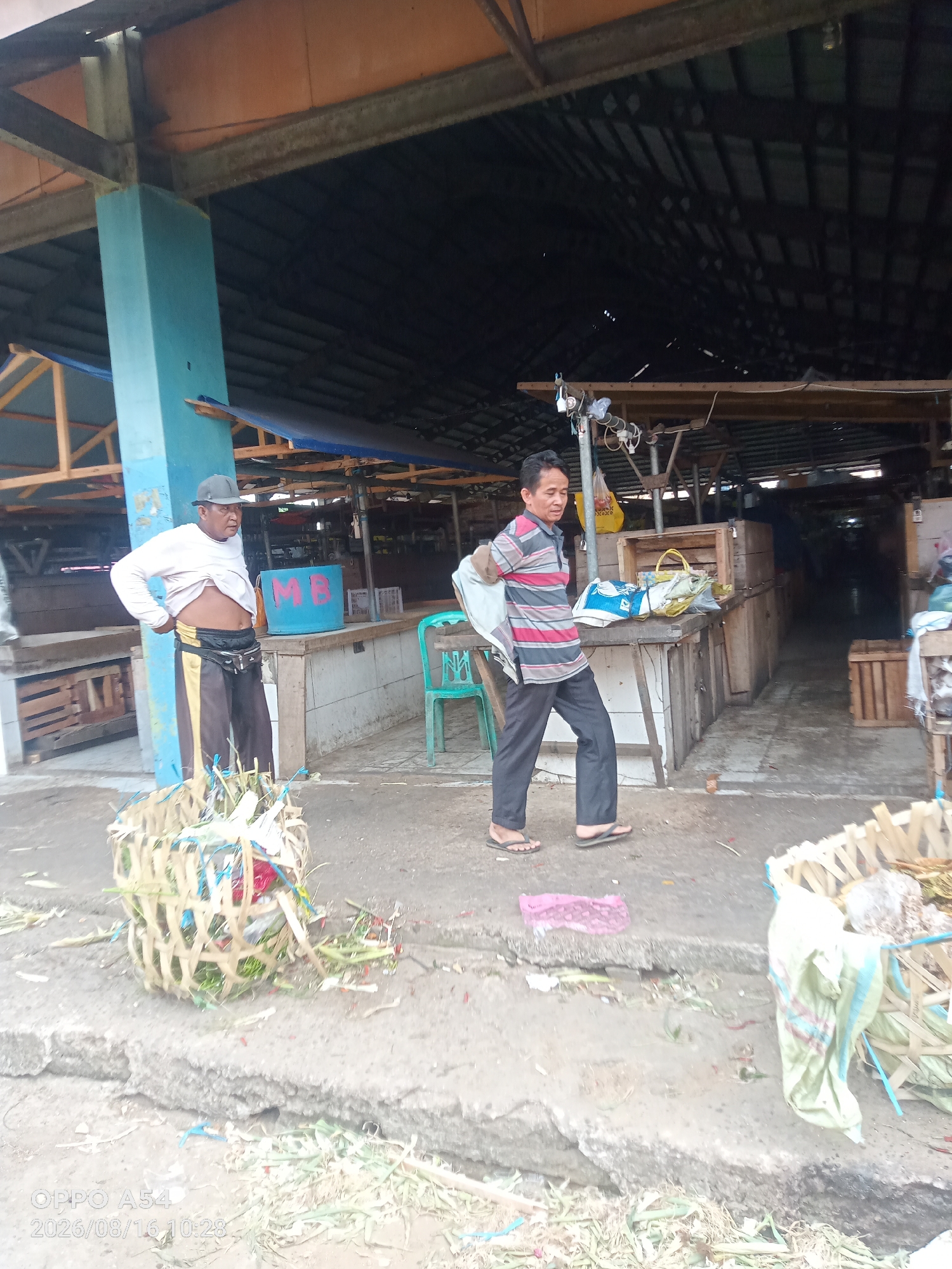 The image depicts an outdoor market scene in Jambi, Indonesia. Two men are present. One man, wearing a grey baseball cap, a light-colored long-sleeved shirt pulled up to reveal his abdomen, and black and gold striped pants, stands next to a blue barrel with "MB" written on it. He is facing away from the camera. In front of him is a woven basket filled with green foliage and some red items. The second man, wearing a horizontally striped t-shirt and dark pants, is walking away from the camera, carrying a white cloth bag over his shoulder. He is wearing flip-flops. Behind the men, there are market stalls with wooden structures and canopies, some appearing to be empty. Baskets and crates are visible within the market stalls. The ground is paved and appears somewhat wet and dirty, with scattered green debris. The overall environment suggests a functional, perhaps slightly disorganized, market.