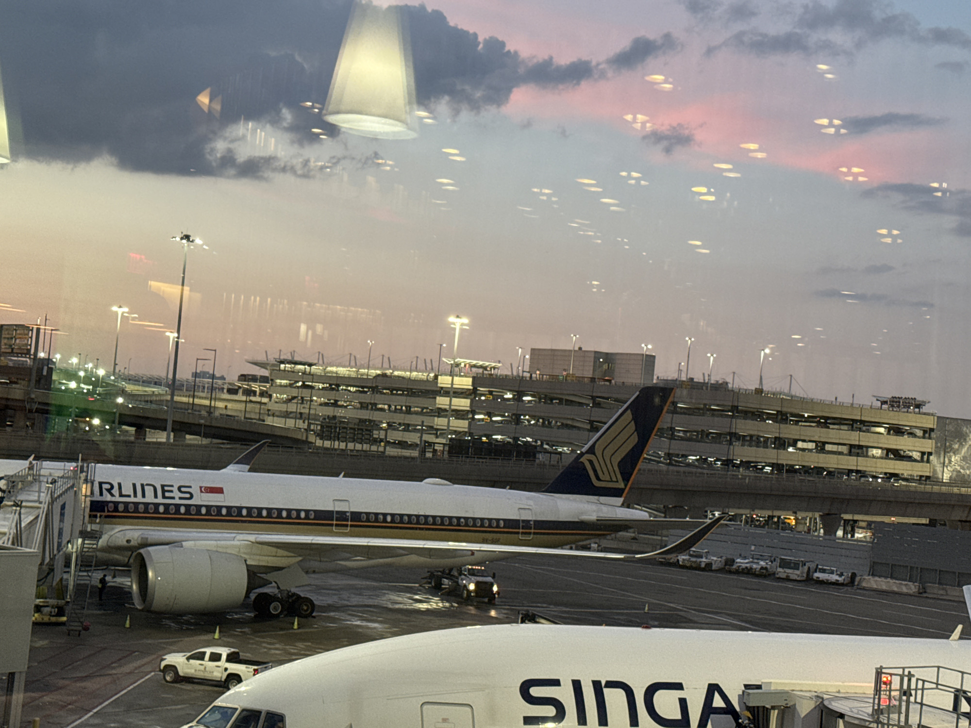 The image captures an airport scene at dusk, with a view through a window that reflects the sky and interior lights. In the foreground, two Singapore Airlines airplanes are visible. The closest airplane has the word "SINGA" printed on its side. The airplane further back is docked at a jet bridge and has the full "AIRLINES" visible, along with the Singapore Airlines logo on its tail. Ground vehicles, including a white pickup truck and baggage tugs, are present on the tarmac. In the background, a multi-story parking garage or terminal building is illuminated, and airport lighting poles are scattered across the scene. The sky displays a gradient of colors, transitioning from a soft pink and orange hue near the horizon to a deeper blue and gray overhead, with scattered clouds. The reflections on the window add a layer of visual complexity, showing abstract patterns of light and dark. The overall impression is one of an active airport during the transition from day to night.