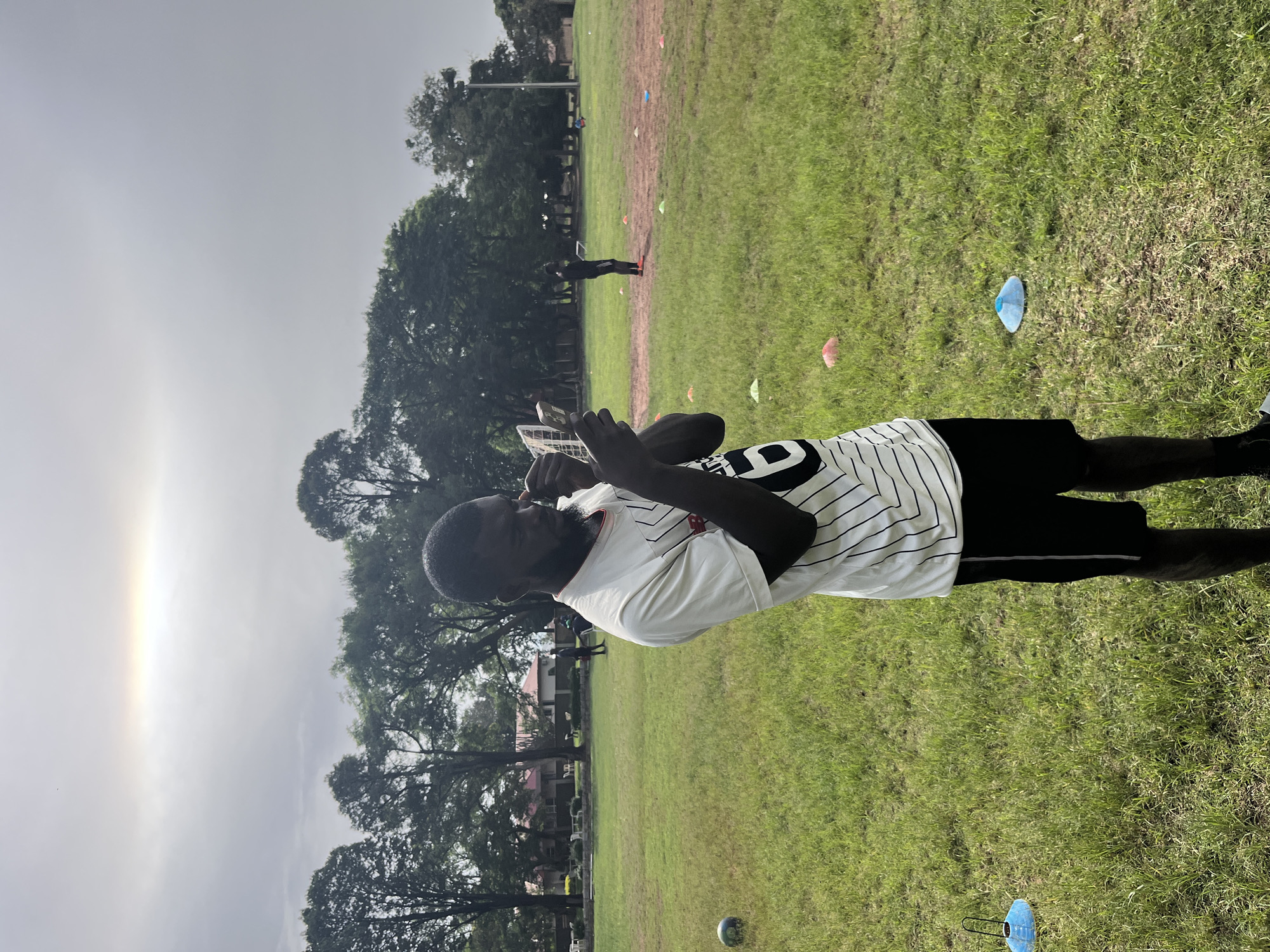 The image depicts a young man standing in a grassy field, possibly a sports training ground, as indicated by the presence of cones and a soccer ball. The man is wearing a white t-shirt with black stripes and black shorts. He is holding what appears to be a compact mirror and is applying something to his face, possibly makeup or sunscreen. In the background, several trees are visible, as well as some buildings. The sky is overcast with a faint rainbow or halo visible. The scene appears to be set in Jos, Nigeria, given the context provided. There are other individuals in the background, some of whom appear to be training.