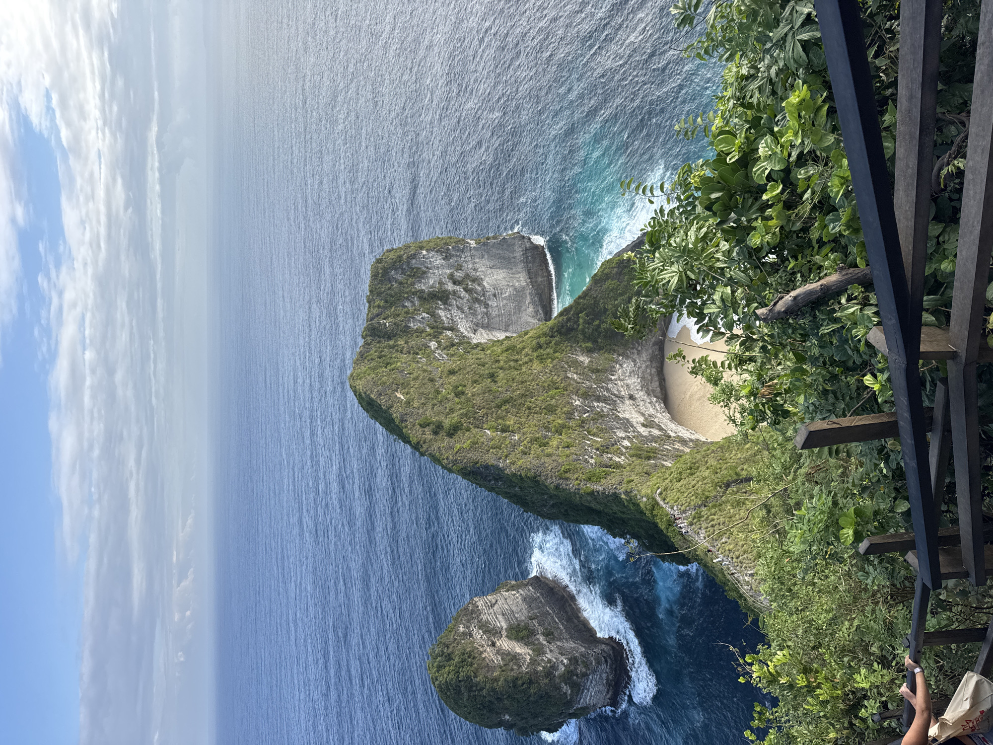 This image depicts a stunning coastal landscape in Nusa Penida, Indonesia, specifically the iconic viewpoint overlooking Kelingking Beach. The foreground features a weathered wooden railing and lush green foliage, with a section of wooden stairs visible, indicating a pathway for visitors. A person's hand and a shopping bag are visible on the left side of the stairs, suggesting recent activity.
Dominating the mid-ground are two prominent rock formations jutting out of the deep blue ocean. The larger formation, shaped like a Tyrannosaurus Rex, is covered in vibrant green vegetation, with steep, layered rock faces. A small, sandy cove is nestled at its base, accessible by a narrow, winding path visible on the cliff face. The smaller rock formation to the left is also vegetated and surrounded by white-capped waves. The turquoise and deep blue waters of the ocean stretch out to the horizon, meeting a soft blue sky filled with scattered clouds. The overall scene conveys a sense of natural beauty, ruggedness, and a popular tourist destination.