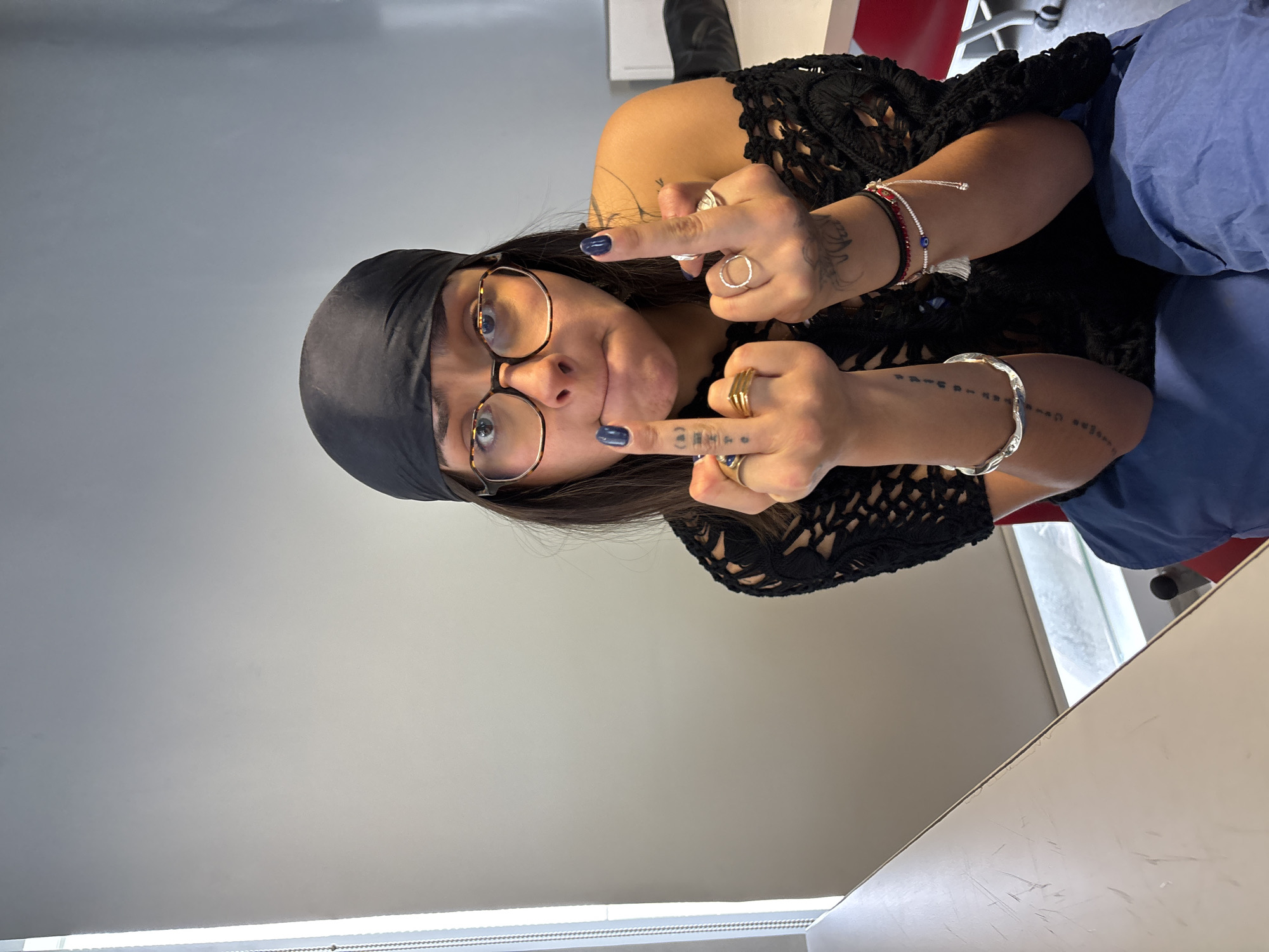 An indoor portrait-style photograph shows a young adult woman making a defiant gesture, extending both middle fingers towards the viewer. She has dark hair styled under a black headscarf or bandana and wears distinctive tortoise-shell framed glasses. Her lips are slightly pursed, conveying a playful yet rebellious expression. Her fingernails are painted a dark blue.
The woman is adorned with several accessories: multiple rings on her fingers, including a stack of gold-toned rings on her right hand and a silver-toned ring on her left. On her wrists, she wears various bracelets, notably a beaded red and white one with an evil eye charm, and a silver-toned cuff bracelet. Tattoos are visible on her fingers, with the letters "GET" clearly visible on the middle finger of her right hand, and other symbols on her left middle finger. Additional tattoos, appearing as lines of small symbols or text, are visible along both forearms.
She is wearing a black, open-knit or crocheted top and blue pants. The background consists primarily of a plain light grey wall, with parts of a red chair and a white table or desk visible on the right side. The lighting is even, indicating an indoor setting. The photograph was taken in Mexico City, Mexico.