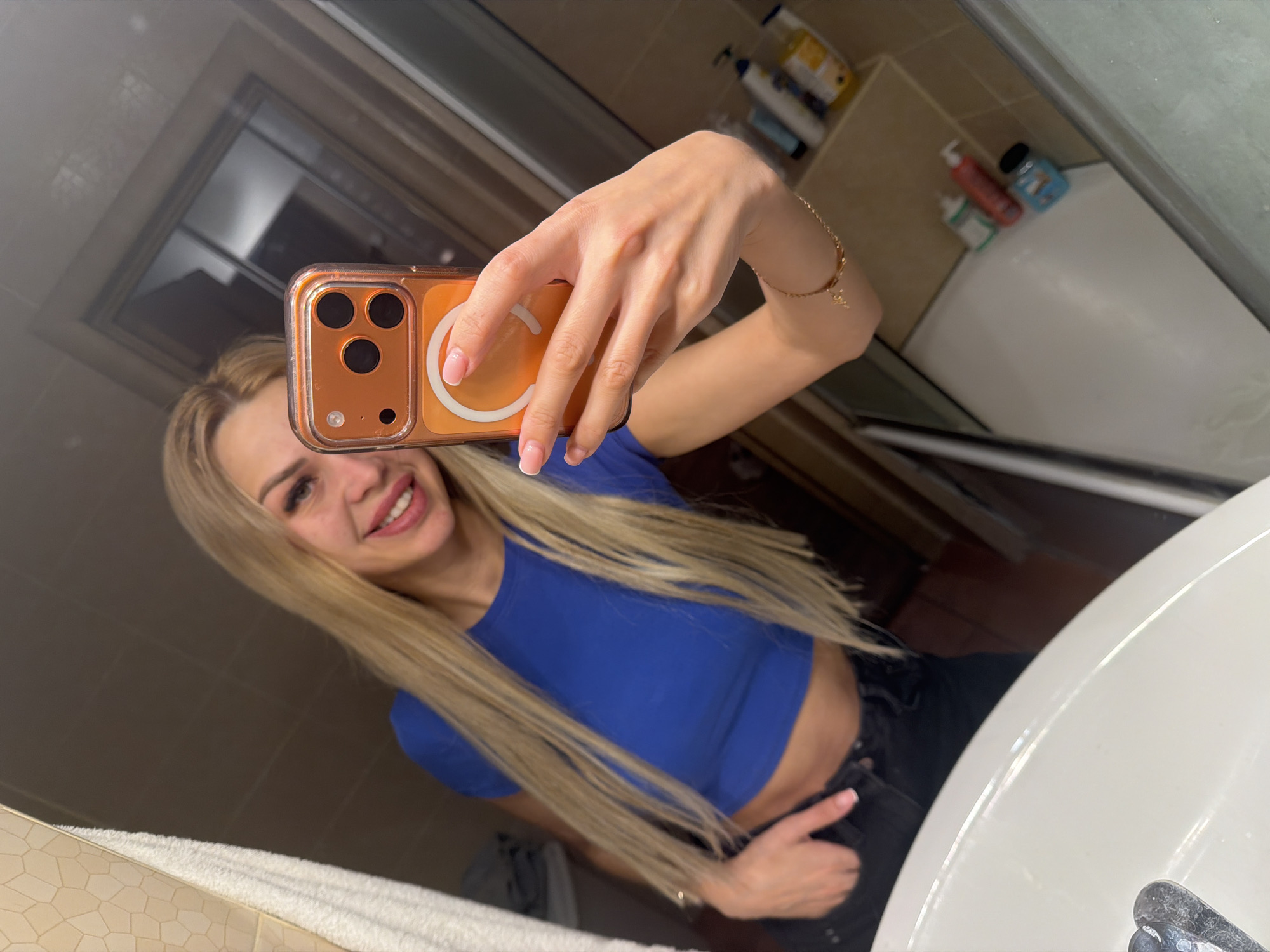 The image shows a young woman with long blonde hair taking a selfie in a bathroom. She is wearing a blue crop top and black jeans, with her hand in her pocket. She is smiling and holding a phone with an orange case. In the background, there is a sink, a towel, and various toiletries. The bathroom appears to be modern and well-lit. The image was taken in Kyiv, Ukraine.