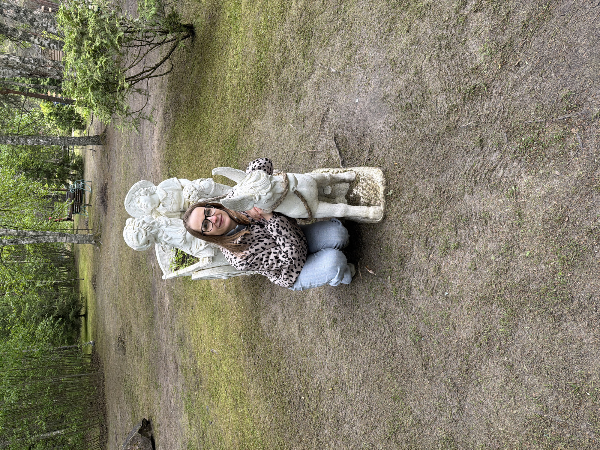 A medium-shot image taken outdoors shows a woman squatting beside a white statue of children holding a donkey on a patch of grass. The woman wears glasses, a brown and black spotted jacket, and blue jeans. The statue is roughly hewn and appears aged. The background consists of grassy areas interspersed with patches of dirt and trees. The location is Zaplav'e, Russia.