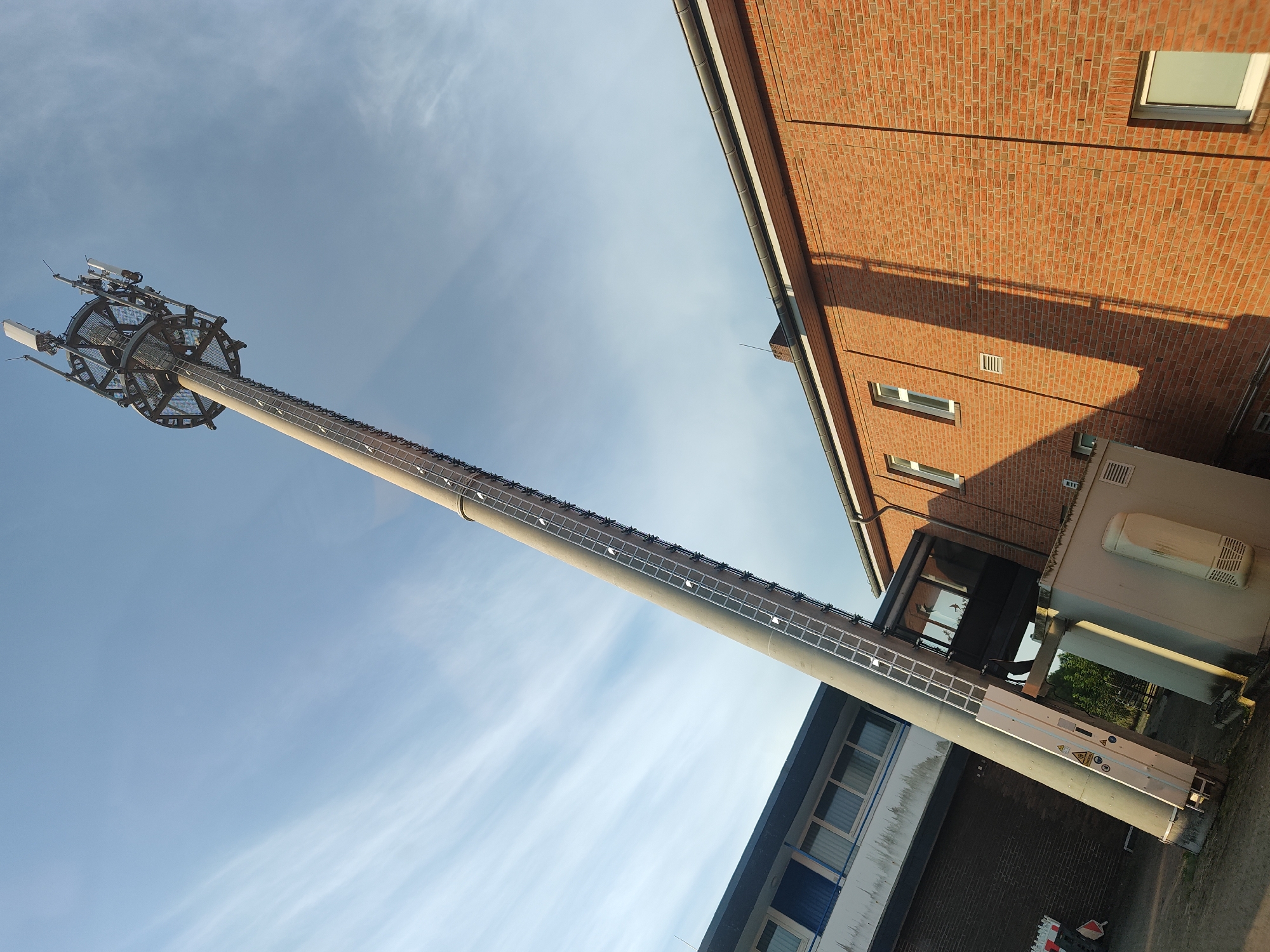 A tall, grey cell tower is the central subject of this image, tilted diagonally from the bottom left to the top right. The tower has a wide base and narrows towards the top, where it supports a circular platform with antennas. A ladder runs vertically along the side of the tower.
To the left of the cell tower, there is a low-rise building with a dark brick base and a lighter upper section featuring windows and blue accents.
To the right of the cell tower is a brick building with a prominent red brick facade. This building has several windows, some of which are dark. A shadow from the cell tower stretches across the brick wall.
At the base of the cell tower, there is a white, box-like structure with vents, possibly housing equipment. The ground in the foreground appears to be asphalt or gravel.
The sky is blue with wispy white clouds, suggesting daylight. The scene is outdoors, likely in an industrial or commercial area of Oldenburg in Holstein, Germany, given the architecture and the presence of a cell tower. There are no people visible in the image.