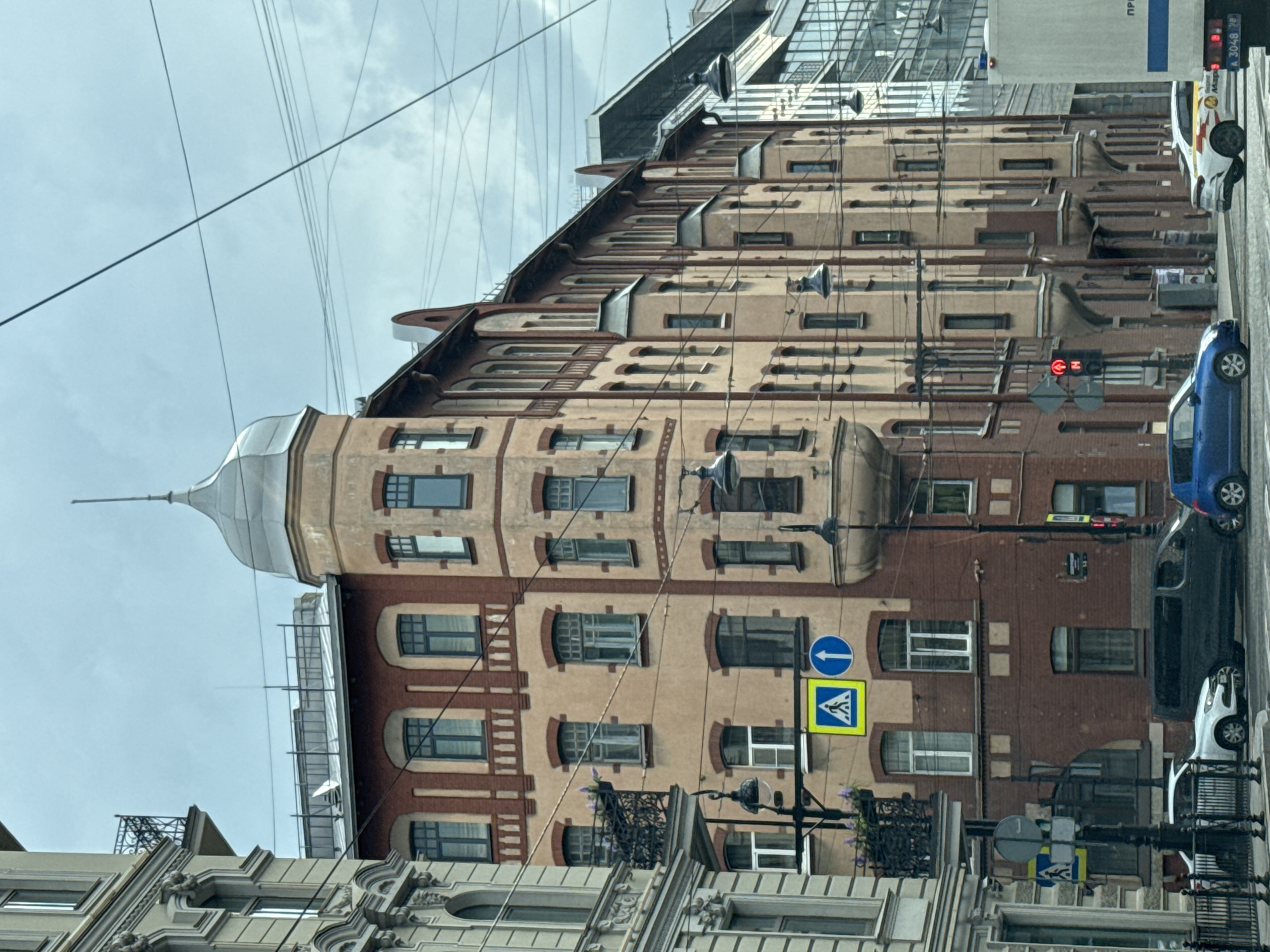 The image depicts a street scene in Saint Petersburg, Russia. A multi-story building with ornate architecture and a distinctive turret occupies the center of the frame. The building's facade is a mix of light brown and reddish-brown bricks, with multiple windows and decorative elements. To the left, another building with similar architectural style is partially visible. The street in front of the buildings has several cars, including a white sedan, a dark minivan, and a blue hatchback, indicating normal city traffic. Traffic signals and pedestrian crossing signs are also present, suggesting an active urban environment. Overhead, a complex network of electrical wires crisscrosses the sky, characteristic of older European cityscapes. The sky is overcast, contributing to the muted lighting of the scene. The overall impression is of a well-preserved historical district within a bustling city.