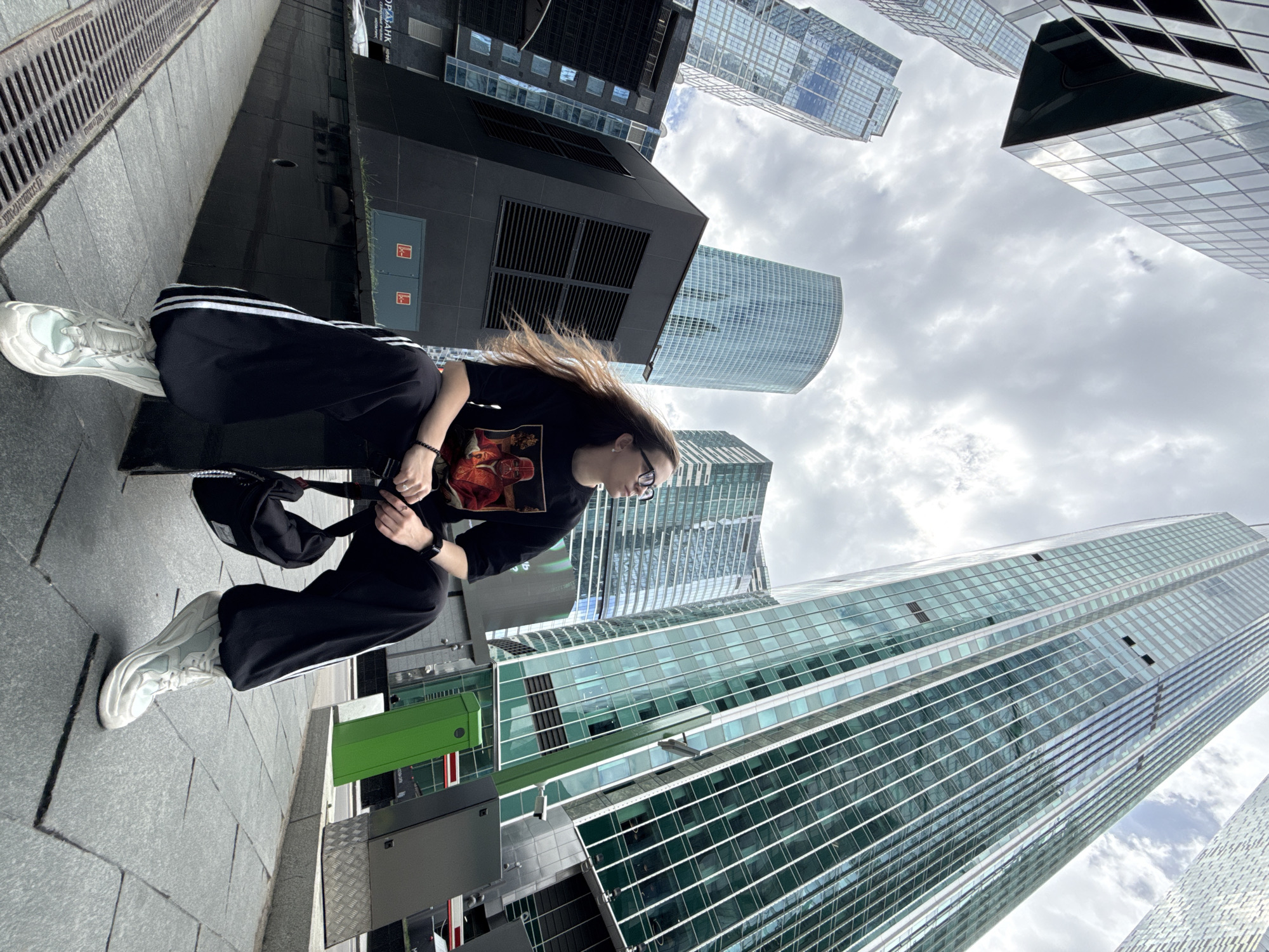 A low-angle shot captures a woman seated on a ledge in an urban setting in Khimki, Russia. Her legs extend to the left side of the frame; she wears sneakers, black pants with white stripes down the sides, and a black t-shirt. She has brown hair and wears glasses. The woman is seated on a dark-colored, flat surface, possibly a bench or ledge. A black bag rests near her. Behind her, a modern cityscape unfolds with tall, glass skyscrapers reaching towards a cloudy sky. The buildings feature a grid-like pattern of windows and reflect the sky. A drain or grate is visible on the left side of the frame.