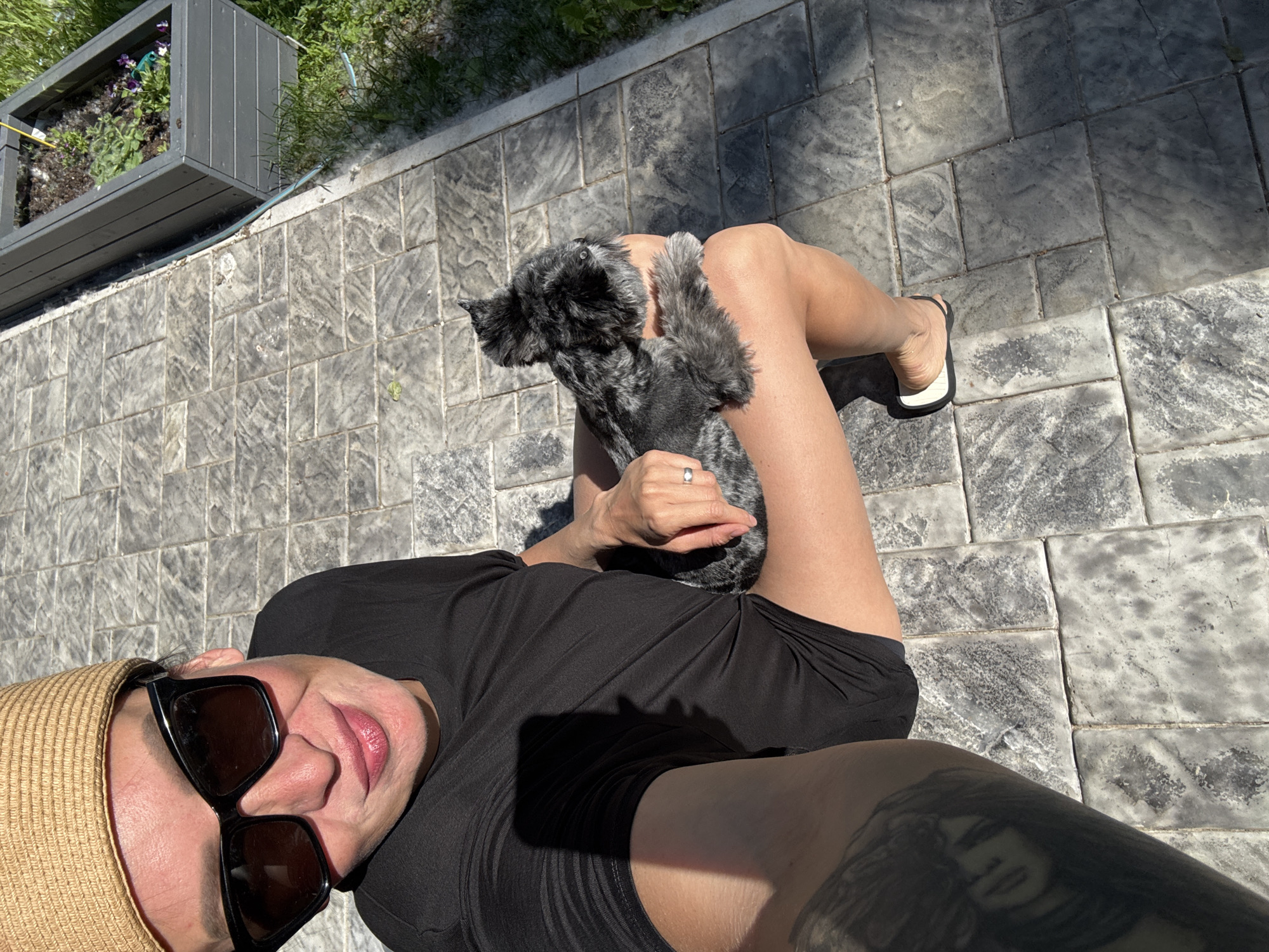 A woman wearing a straw hat and sunglasses sits on a stone-paved patio in Chaikovsky, Russia. She is holding a small, black and gray dog. The woman is wearing a black t-shirt and has a large tattoo on her left arm. A planter box filled with plants is visible in the background. The scene is illuminated by bright sunlight.
