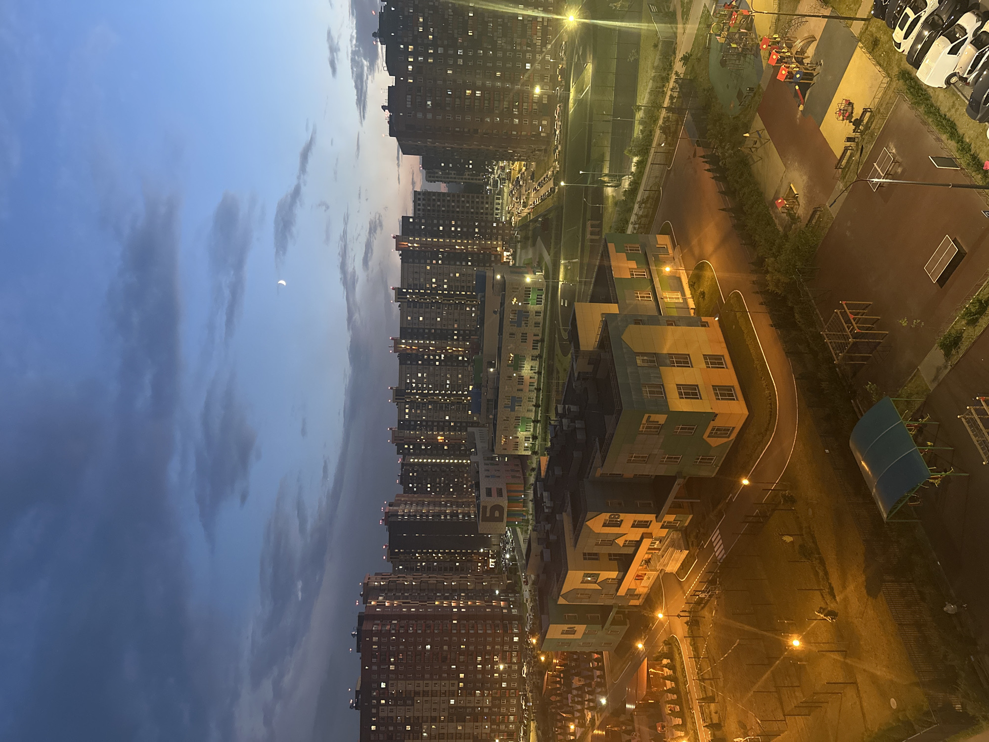This media file depicts a twilight scene in a residential area, likely in Mirny, Russia. The primary subjects are numerous apartment buildings, characterized by their uniform, multi-story structures. Many windows are illuminated, suggesting the evening hour and the presence of inhabitants.
The setting includes outdoor recreational spaces such as playgrounds with colorful equipment and sports courts, some of which are lit by streetlights. Rows of parked cars are visible along the streets and in parking areas, indicating a populated neighborhood. There are also smaller, modern buildings with colorful facades, possibly schools or community centers.
The time of day is clearly evening, as indicated by the deep blue of the sky and the artificial lighting illuminating the scene. The sky contains some wispy clouds, and a sliver of the moon is visible. The overall mood is tranquil and urban. No specific event is actively taking place, rather it's a snapshot of daily life in a modern housing development.
The image is oriented vertically, requiring rotation for a standard view. The architecture suggests a contemporary urban environment. No legible text is visible within the frame.