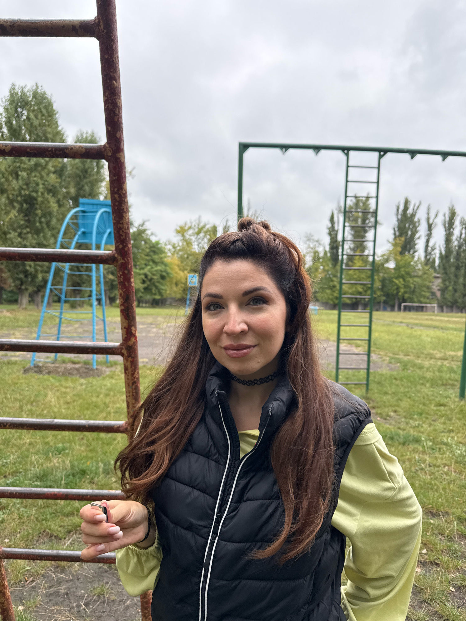 A woman with long brown hair styled in a half-up, half-down bun stands in front of a rusted metal playground structure. She is wearing a black puffer vest over a light green long-sleeved shirt and a black choker necklace. Her right hand is visible, holding a silver object. In the background, a blue metal playground slide and green monkey bars are visible, along with trees and a cloudy sky. The scene appears to be an outdoor recreational area, possibly a schoolyard or public park, in Kryvyi Rih, Ukraine.