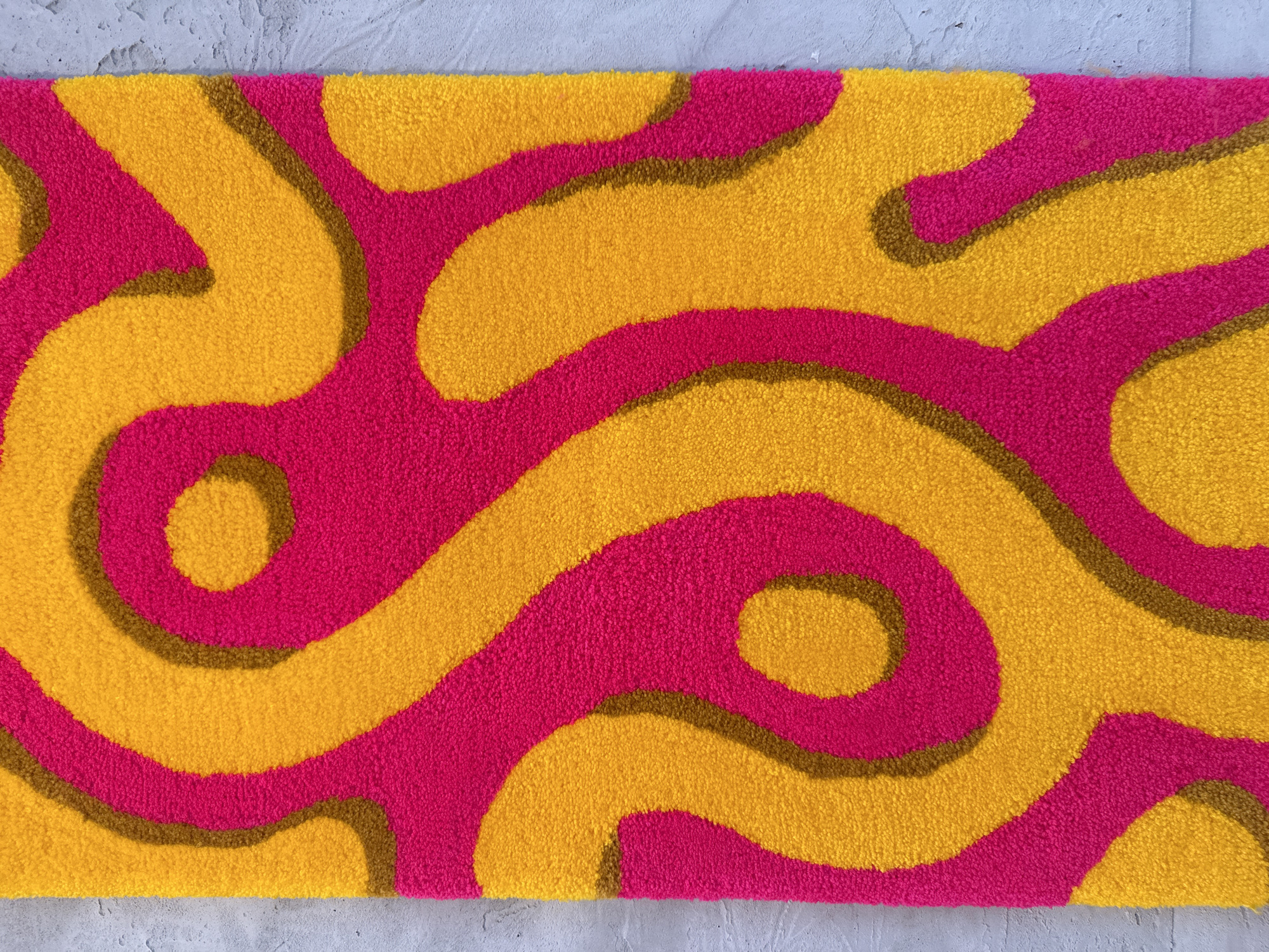 The image depicts a close-up view of a rectangular shag rug with an abstract, curvilinear pattern. The rug is primarily composed of two distinct colors: a vibrant, hot pink and a saturated golden yellow. A thin, dark brown outline traces the edges where the pink and yellow fields meet, creating definition between the shapes. The pile of the rug appears thick and plush, suggesting a soft texture. The rug is positioned on a textured, light gray surface, possibly concrete or a similar material. The scene does not contain any people or indication of a specific location in Budva, Montenegro.
