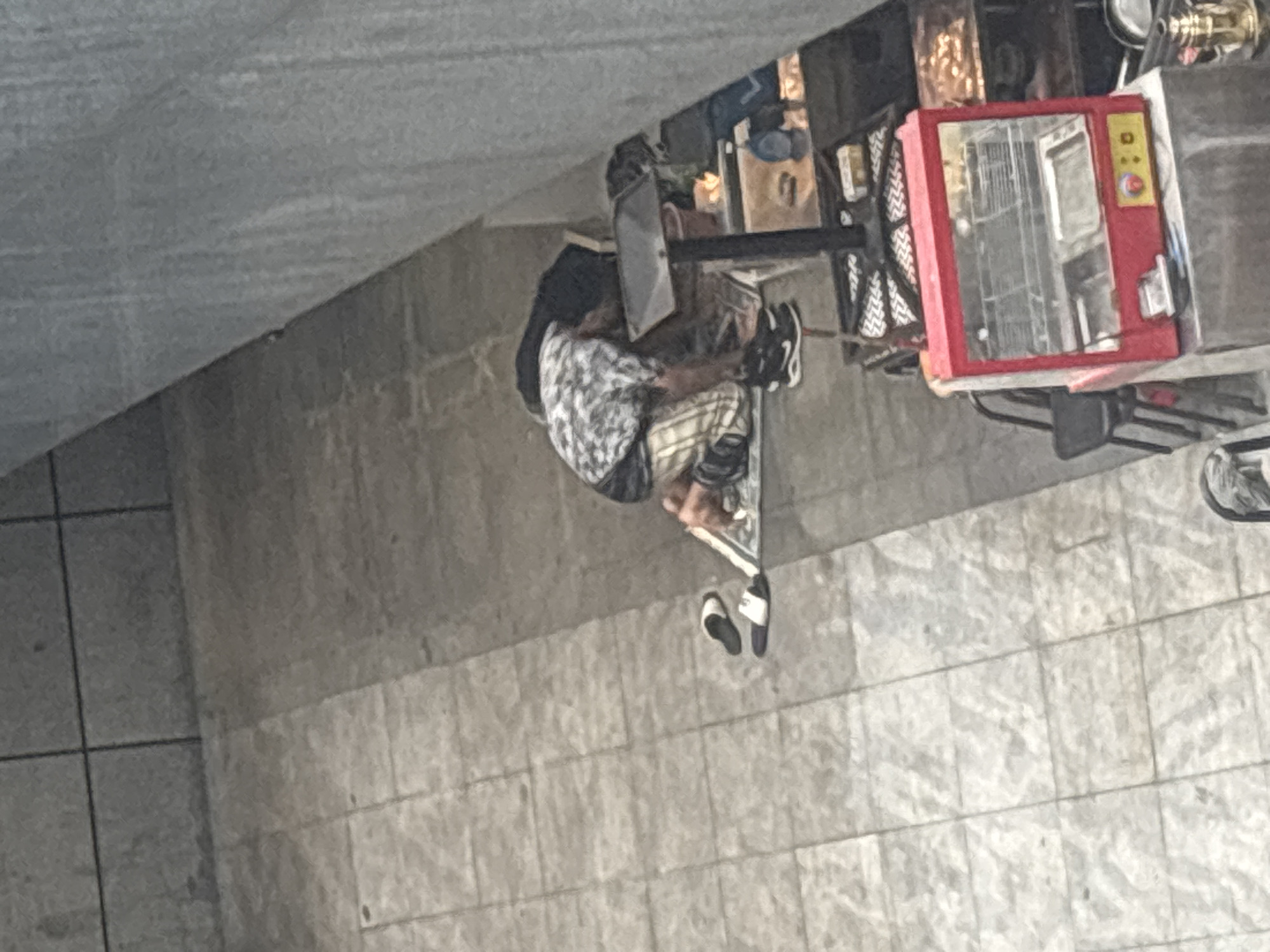 A person is kneeling on a tiled floor in what appears to be an outdoor urban setting. They are wearing a patterned shirt, plaid shorts, and black athletic shoes. To their left are two white and black slides. The person is reaching towards a black stand with a reflective surface, near which is a red food warming display case. The background shows a textured wall and more tiling. The location is identified as Liwan District, China.