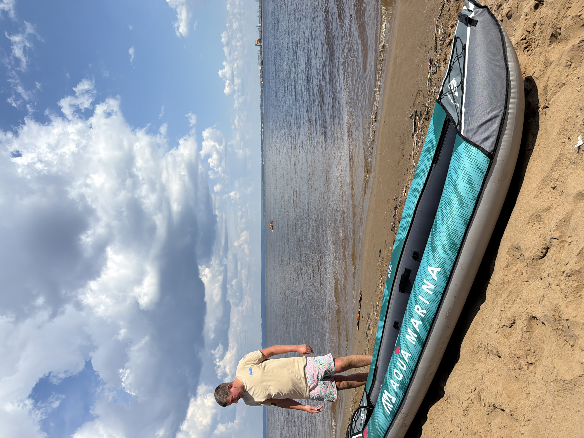 An adult male wearing a light-colored t-shirt and patterned shorts stands on a sandy beach next to an inflatable kayak. The kayak is teal and gray with "AQUA MARINA" written on it. The man is looking down at the kayak. The body of water in the background is calm with a distant shoreline and some industrial structures. The sky is partly cloudy. The overall scene suggests an outdoor recreational activity in a waterfront location. The location is identified as Noviy, Russia.
