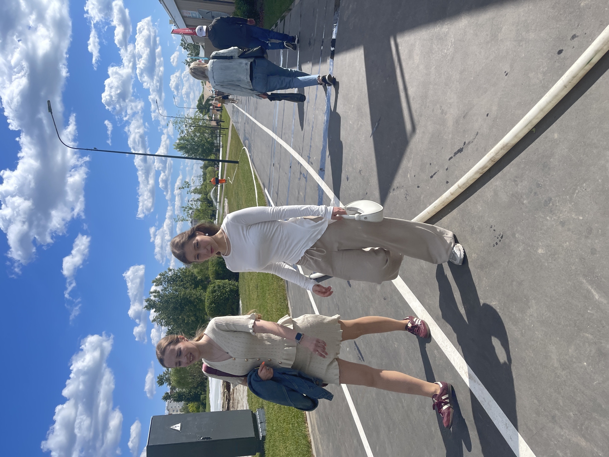 Two young women are walking on a paved path outdoors. The woman on the left wears a light-colored knit top and skirt, with a denim jacket draped over her left arm. She has a maroon strap across her body. The woman on the right wears a long-sleeved white top and loose-fitting tan pants, carrying a white handbag. They are walking towards the right side of the frame. Several other people are visible walking further down the path, some with their backs to the camera. The path is marked with white lines and has a blue accent line along the left edge. The environment appears to be a park or outdoor recreational area with green grass, trees, and bushes. In the background, there are buildings and a cloudy blue sky. This scene is likely in Moscow, Russia, given the context provided by the prompt.