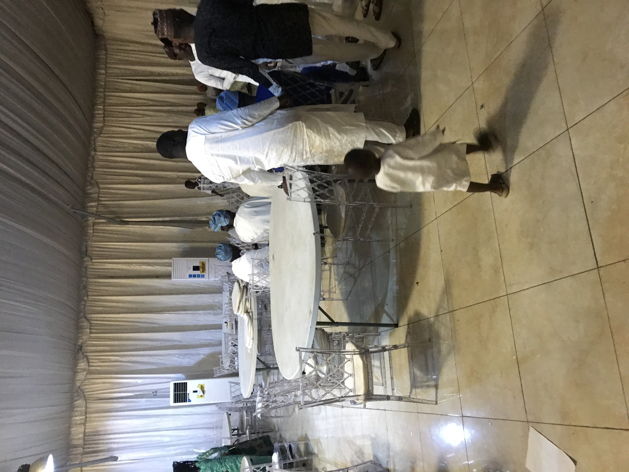 The image shows a gathering of people in what appears to be a tent or large indoor space, possibly in Oko Erin, Nigeria. Several round tables covered with white tablecloths are arranged with clear, decorative chairs. Some individuals are seated around the tables, dressed in traditional attire. One child is walking away from the tables. White drapes line the walls of the tent. There are portable air conditioning units installed on the wall. The floor is tiled.