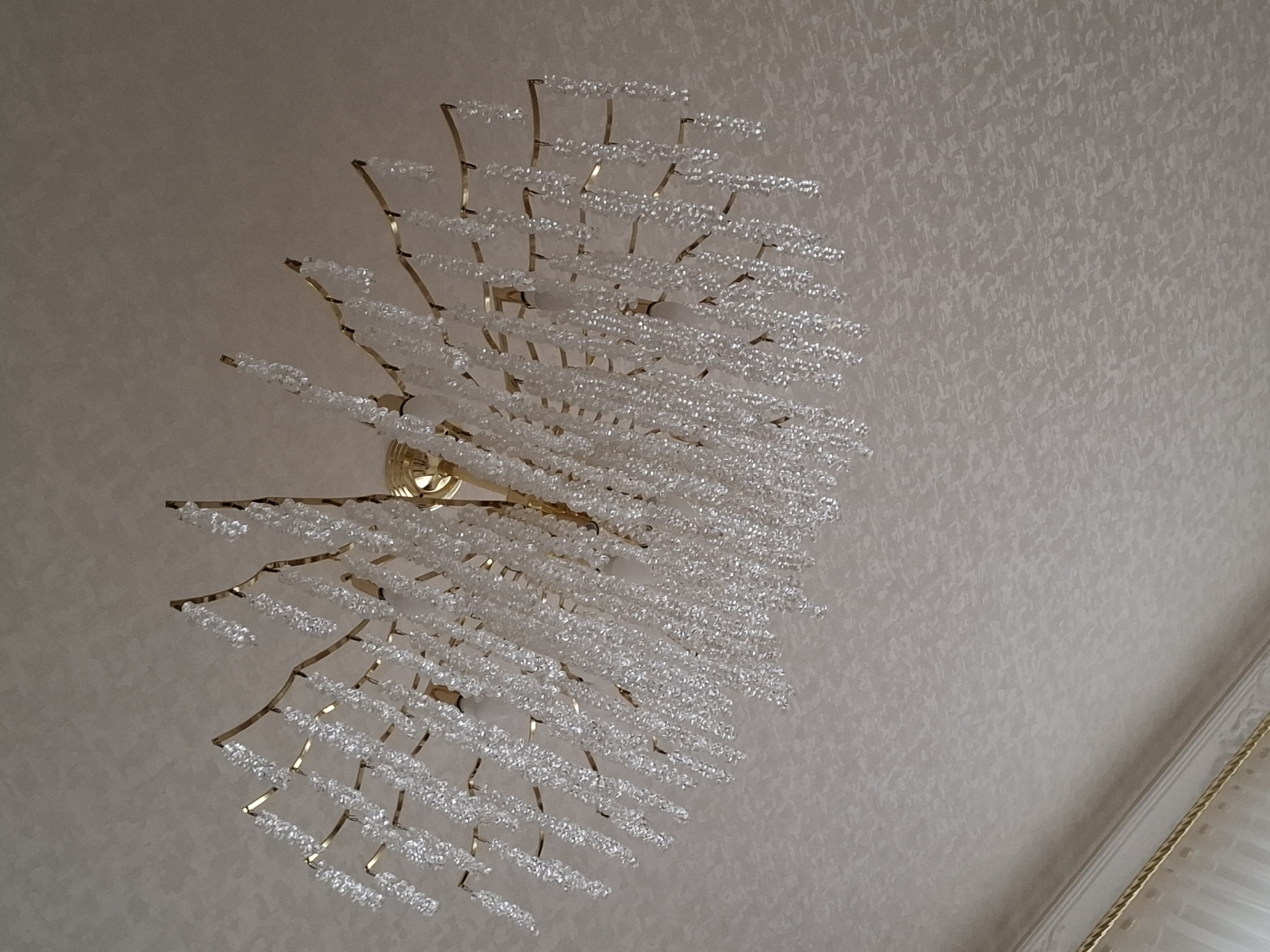 A chandelier hangs from a textured, light-colored ceiling. The chandelier has a gold-colored frame with numerous strings of clear, faceted beads that hang down in various lengths, resembling icicles or fringe. One light bulb is visible within the chandelier structure. The ceiling has a subtle, repeating pattern. In the lower left of the image, a portion of a window treatment with decorative gold trim is visible. There is no information in the image to indicate its location in Urus-Martan, Russia.
