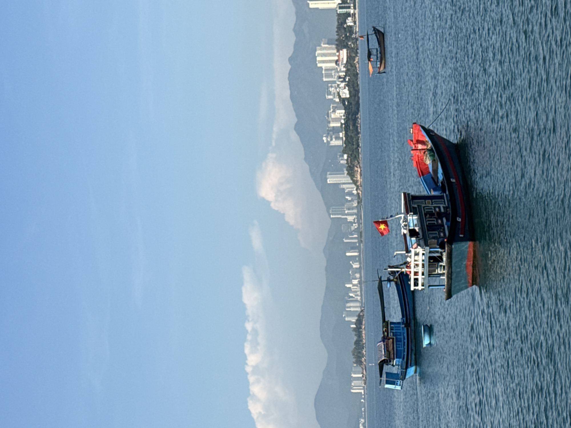 In the city of Nha Trang, Vietnam, a serene seascape unfolds under a clear blue sky dotted with soft white clouds. The foreground is dominated by the tranquil blue waters of the sea, with gentle ripples indicating a calm atmosphere. Several boats are anchored in the water, adding a touch of activity to the scene. A prominent blue and red fishing boat is positioned in the center, featuring a canopy and a Vietnamese flag proudly displayed. To its left, a smaller blue boat floats calmly, and a bright blue bucket is visible near it. Further in the distance, another small boat with an orange canopy is seen. The background reveals a picturesque coastline with lush green trees and a modern cityscape characterized by numerous tall buildings and high-rises, suggesting a vibrant urban area. Majestic mountains form a dramatic backdrop to the city, their peaks shrouded in a light haze. The overall scene captures a blend of natural beauty and urban development, a common characteristic of coastal cities in Vietnam.