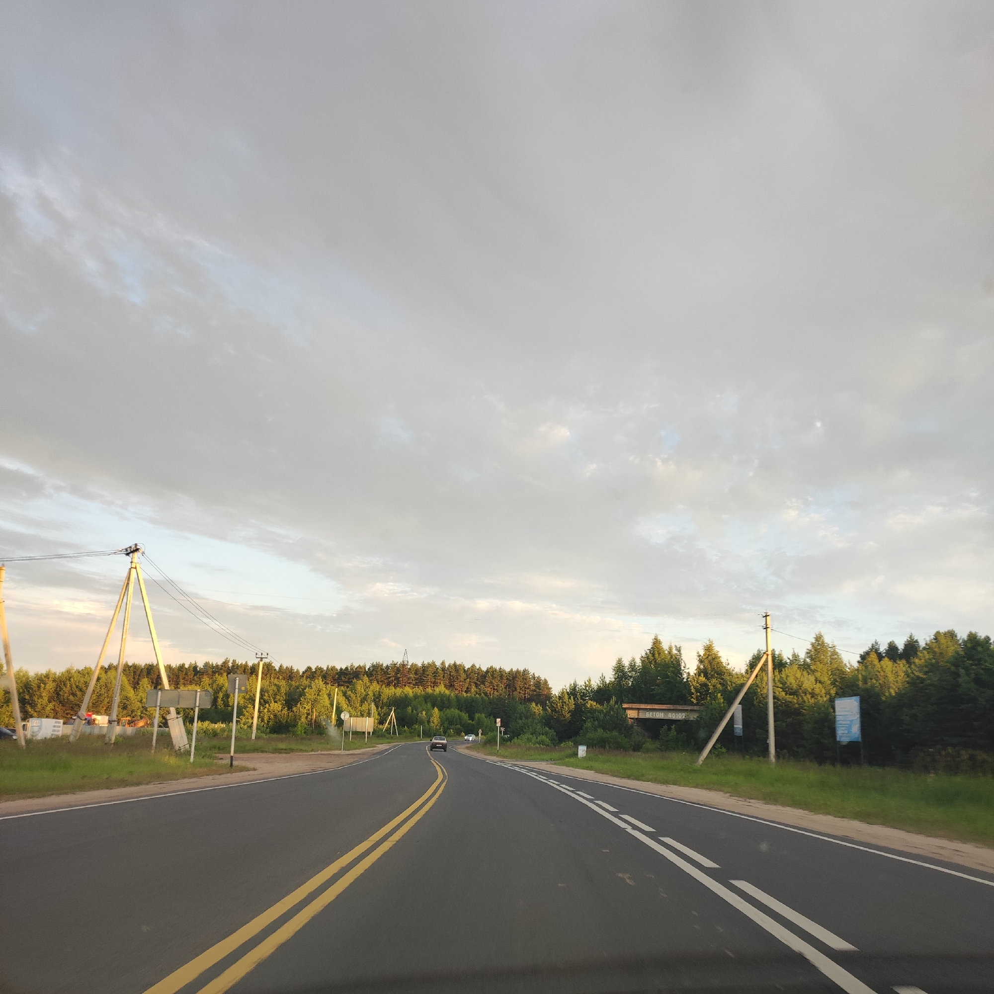 The image captures a rural road scene at dusk, with a wide asphalt road curving to the left. The road is marked with a double yellow line in the center and a white dashed line on the right shoulder. On the left side of the road, there are power lines supported by tall, triangular wooden poles, and a small collection of parked vehicles including a white van with red flashing lights, and what appears to be construction equipment. Beyond the road and to the left and center, a dense forest of green trees forms a backdrop, with a clear sky with wispy clouds above. On the right side of the road, there is a sign with the Cyrillic text "БЕТОН 40107" and another blue sign with text, all set against the forest. A dark sedan and a lighter colored car are visible driving on the road in the distance. The overall impression is one of a peaceful, somewhat isolated, rural setting.