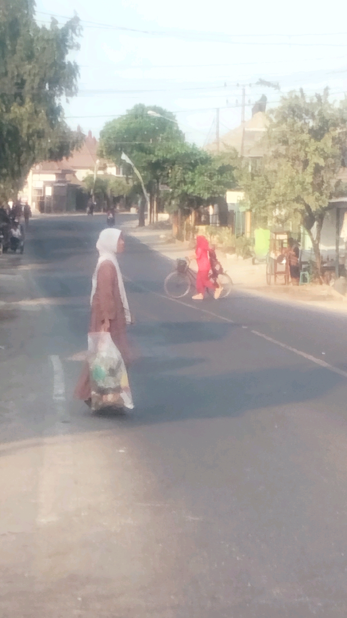 In a city street in Palang, Indonesia, a woman in a light brown hijab and dress walks across a road, carrying a plastic bag filled with groceries. She is in the foreground, on a pedestrian crossing marked by white lines. In the middle ground, a person wearing a bright red hijab and outfit rides a bicycle. The road is paved and appears to be moderately busy, with some buildings and trees lining the sides. The sky is bright and hazy, suggesting it may be daytime.