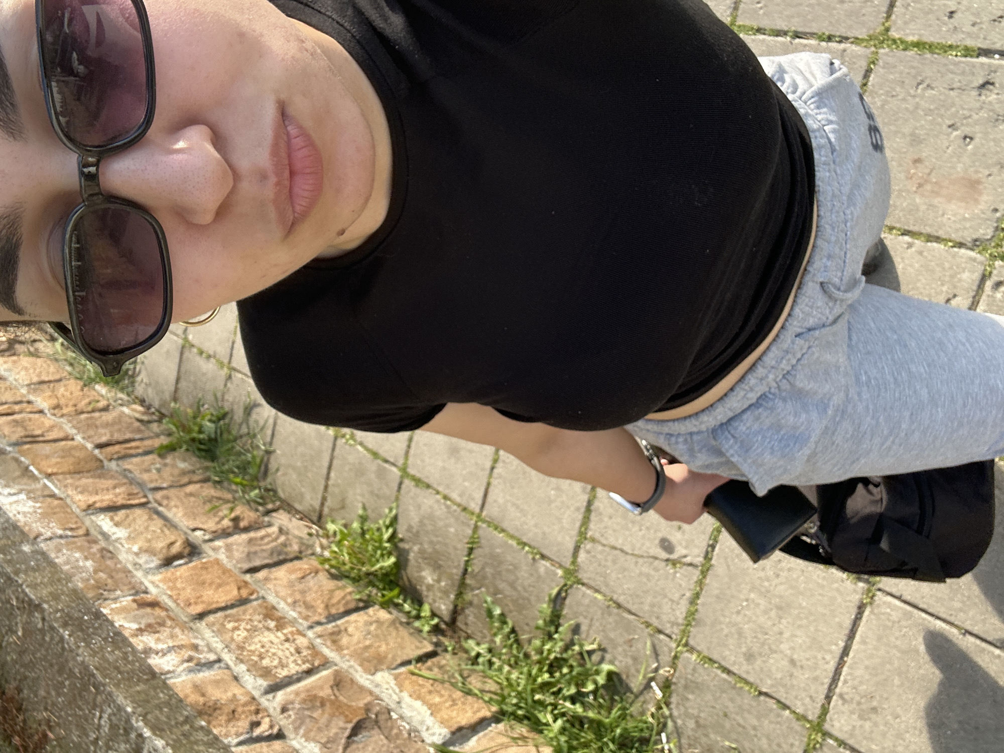 A first-person, overhead perspective captures a person standing outdoors. The individual wears sunglasses, a black t-shirt, and gray sweatpants. A black purse is visible near the person's legs. The background includes brickwork and paving stones with vegetation growing between the cracks. The location is unknown, and there is no information to indicate that this is Burgas, Bulgaria.