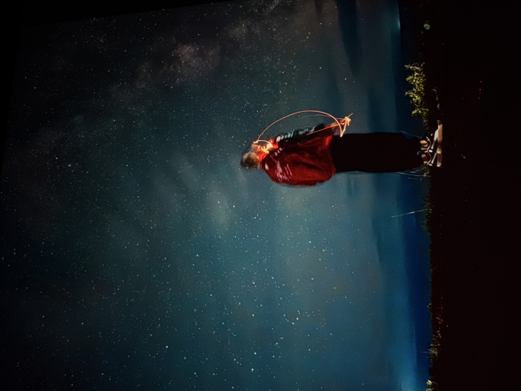 A lone individual, silhouetted against a vast, starry sky, is the central subject of this image. They are positioned in the lower right quadrant, facing away from the viewer and towards the celestial expanse. The person is wearing a dark bottom garment and a bright red jacket with some lighter colored accents. Their arms are raised, and a streak of orange light, indicative of a light painting or sparkler, encircles them, suggesting movement or a performed action.
The background is dominated by the night sky, filled with countless visible stars and the faint, ethereal glow of the Milky Way. The sky transitions from a deep, inky black at the top to a gradient of blues and purples towards the horizon. Below the sky, a dark, silhouetted landscape is visible, featuring a low, undulating terrain with some indistinct vegetation. The ground appears to be covered in dark earth or grass. The overall impression is one of a remote, open area under a clear, dark sky, likely outside of a major urban center, precluding a specific location within Irkutsk, Russia, without further contextual information.