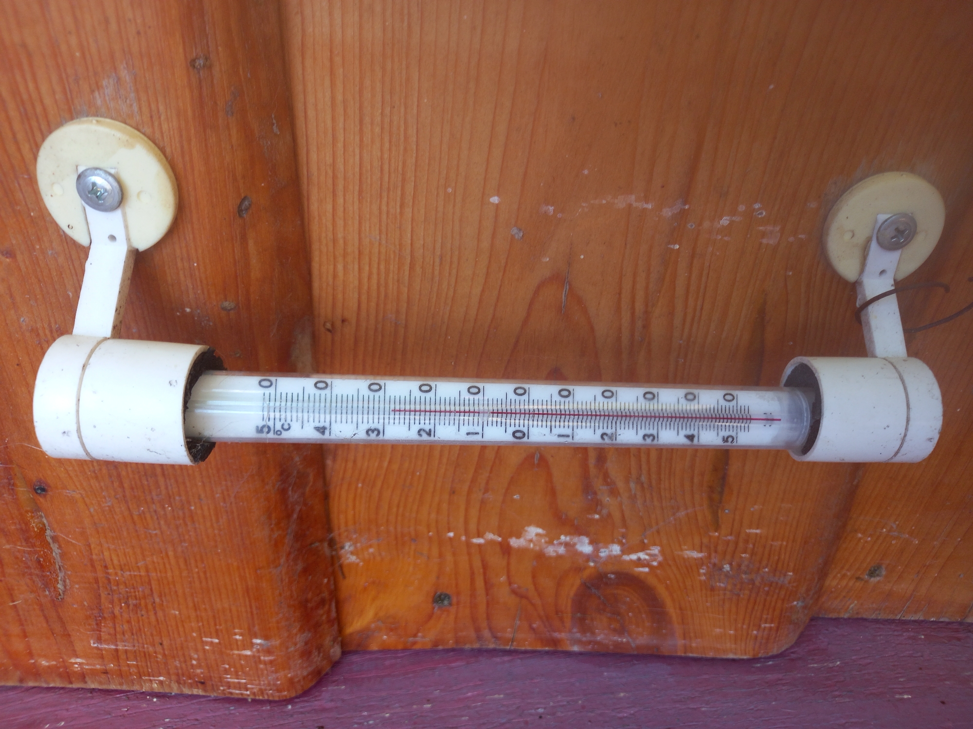 A close-up, high-angle, vertical shot shows a thermometer attached to a wooden wall. The thermometer is white with a glass tube containing a red liquid. The temperature scale is marked in Celsius, with markings for 0, 5, 10, 15, 20, 25, 30, and 35 degrees. The red liquid level is just below the 5-degree mark, indicating a temperature slightly above 4 degrees Celsius. The thermometer is held in place by white plastic brackets that are screwed into the wood. The wooden wall is made of rough-hewn planks with visible grain. The wall appears to be weathered and has some white splotches and dirt. The background is a blurred continuation of the wooden wall, with a hint of purple on the far left edge. The location is Adutiškis, Lithuania.