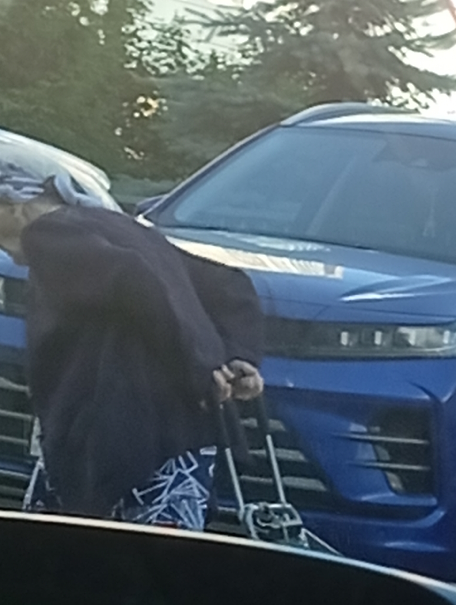A person, appearing to be elderly, is bent over, holding onto a walker. They are wearing a dark jacket and patterned pants. They are standing next to a blue SUV. The background shows trees and foliage. The scene appears to be an outdoor setting, possibly a parking lot or street.