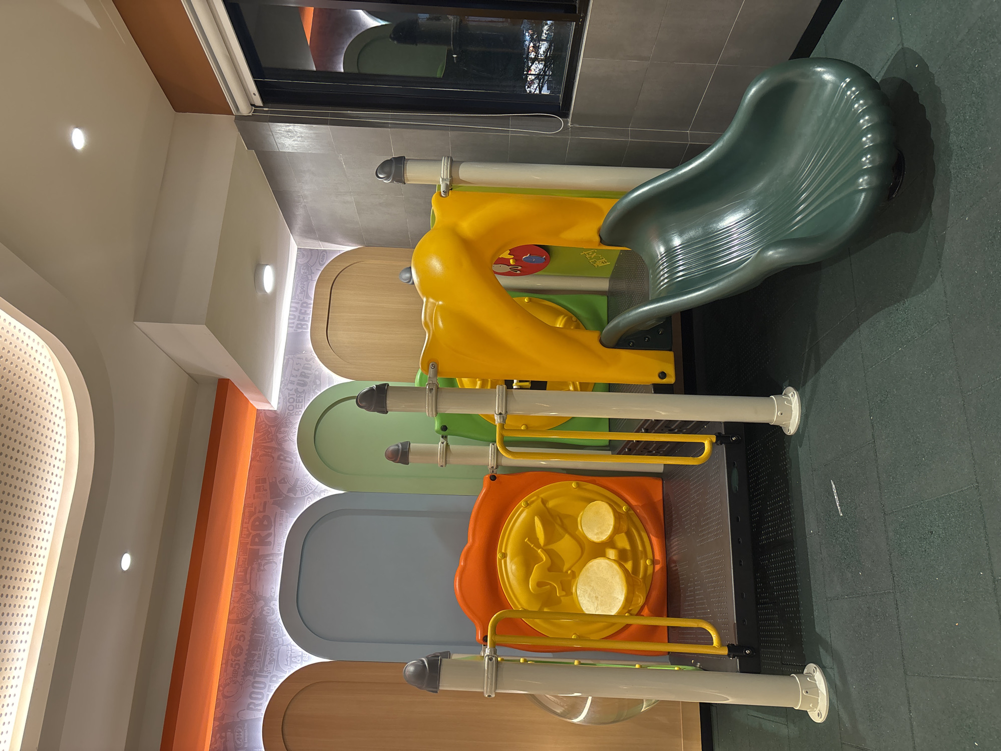 The image depicts an indoor children's playground located in Sukabakti, Indonesia. The playground features a multi-colored play structure with a green slide, yellow and orange platforms, and a red panel with a circular yellow design resembling drums. The structure is supported by white poles. The floor is covered with dark green rubber matting, and shadows are cast from the playground equipment. In the background, there are arched alcoves with wooden, blue, and green panels. The ceiling has a modern design with perforated panels and recessed lighting, and a strip of orange lighting runs along the wall. A window with blinds is visible on the right side, reflecting the interior and some exterior lights. The overall environment appears to be clean and well-maintained, suggesting a commercial establishment like a restaurant or a dedicated play area. There are no people present in the image.