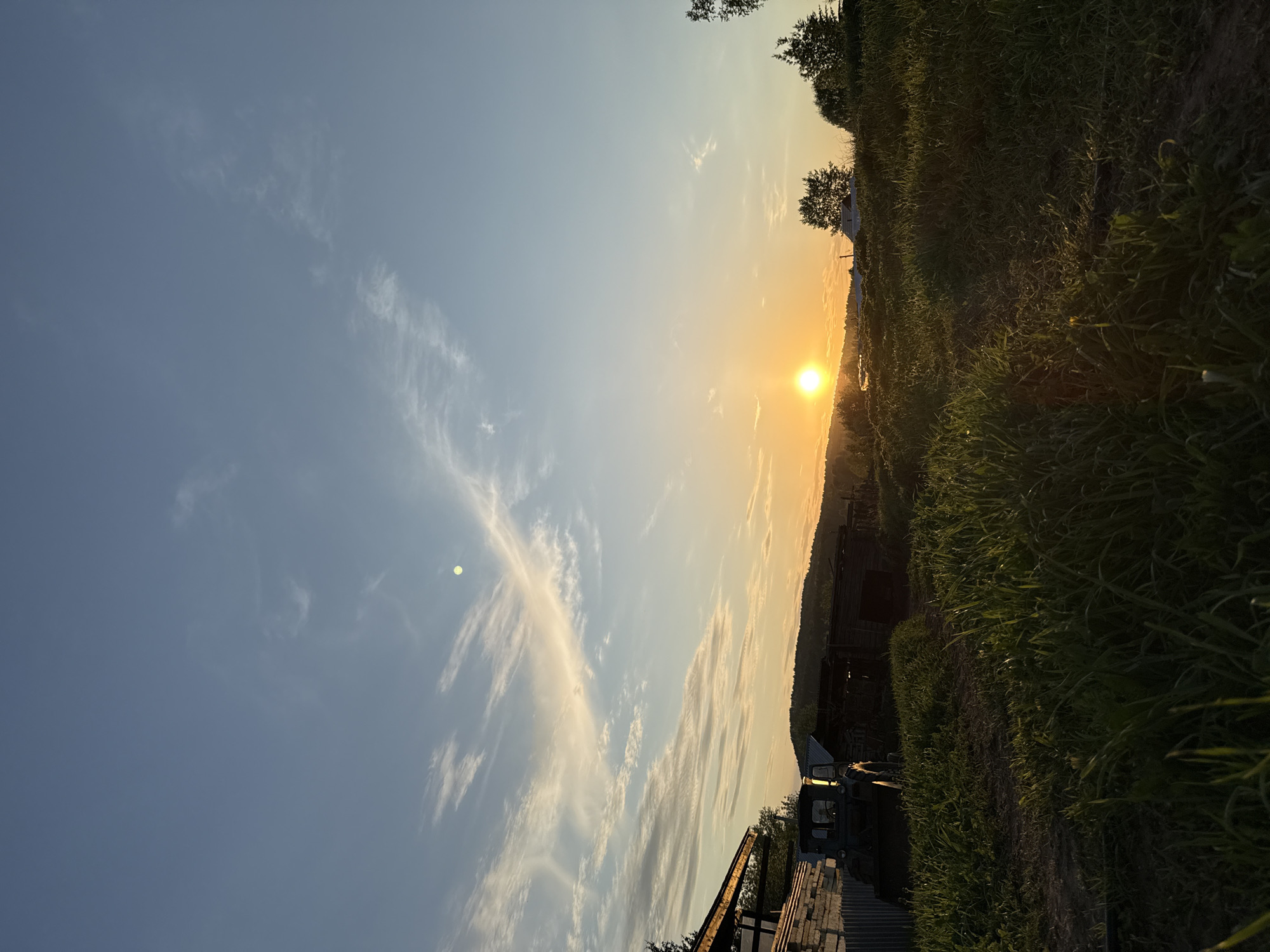 The image depicts a rural scene, likely near the city of Nizhnyaya Chermoda, Russia, at sunset. The sun is positioned on the horizon, casting a warm, golden light. The sky transitions from a clear blue to an orange hue near the horizon. Clouds are sparsely scattered throughout the sky, with some elongated cloud formations stretching across the upper portion of the image.
In the foreground, there is a field or uncultivated land covered with grass and low-lying vegetation. An agricultural vehicle, possibly a tractor or other farm equipment, is situated in the lower part of the frame. Adjacent to the vehicle, there is a stack of concrete blocks or bricks. A dilapidated wooden structure or shed is present in the middle ground, partially obscured by the vegetation. Beyond the structure, a slightly elevated landscape or hillside is visible. The scene suggests a quiet, countryside setting.