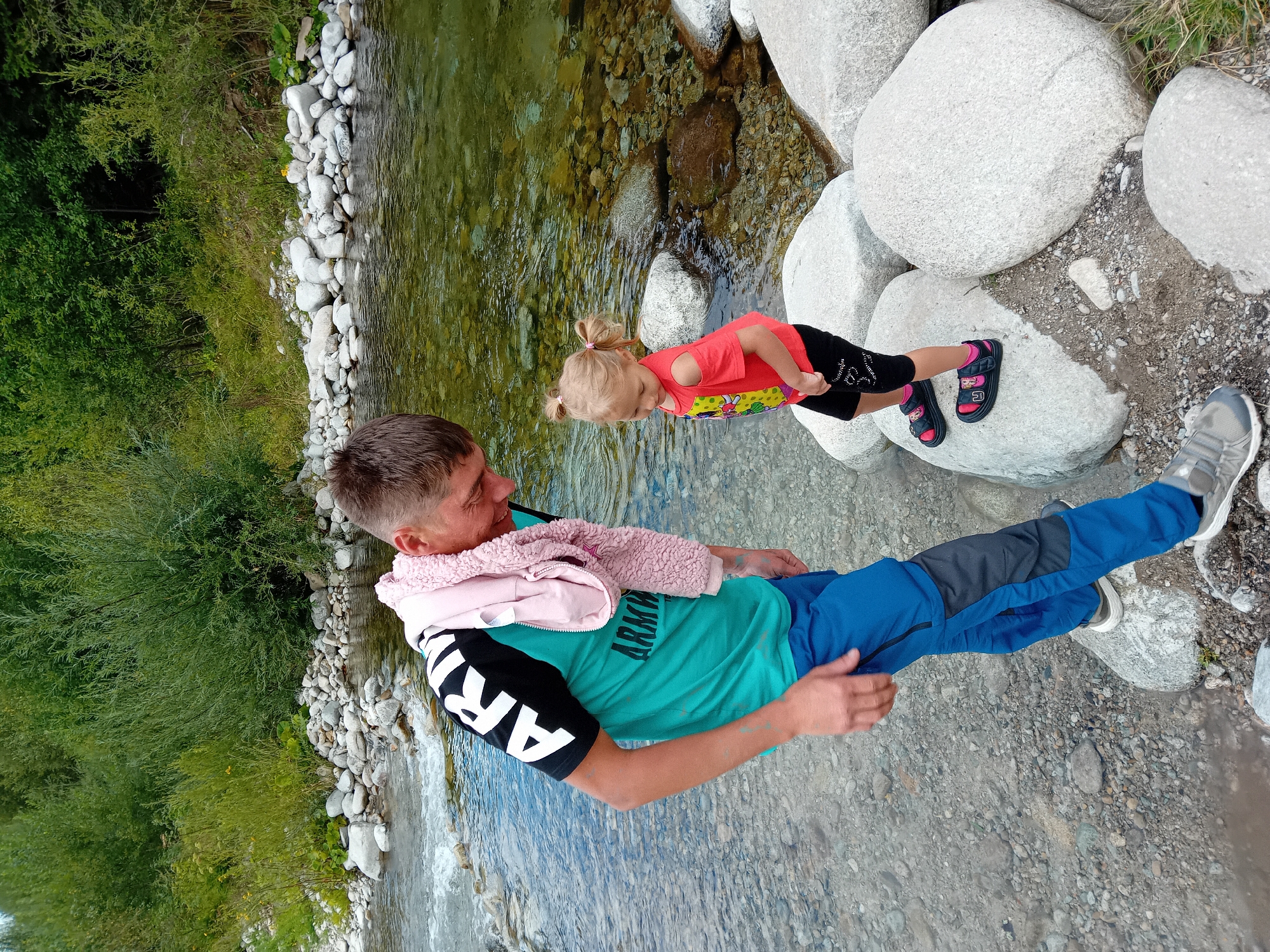 In Zuberec, Slovakia, a man and a young girl stand near a shallow, clear river. The man, wearing a turquoise t-shirt with black and white lettering, blue pants, and grey sneakers, has a pink hooded jacket draped over his shoulders. The girl, dressed in a red t-shirt and black shorts with pink sandals, stands on large, light-colored rocks in the river. The riverbed is visible, composed of small rocks and pebbles. The background features lush green vegetation and trees.
