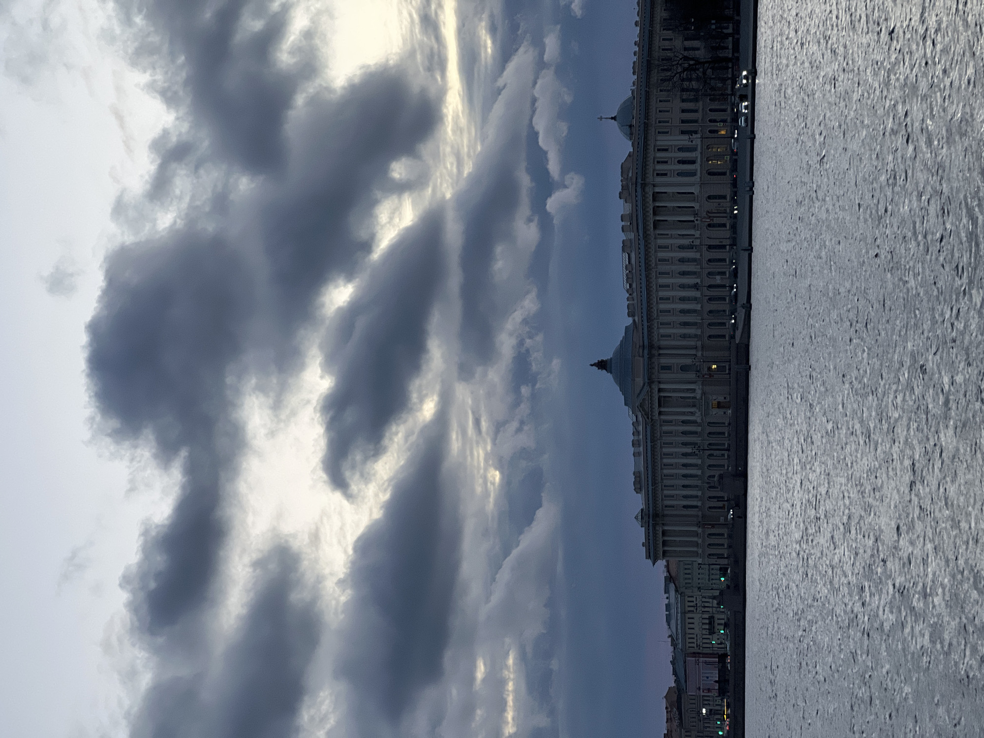 The image depicts a cityscape in Saint Petersburg, Russia, under a cloudy sky. The Neva River dominates the right side of the frame, its surface reflecting the overcast light. Along the riverbank stands the Winter Palace, a Baroque-style building with multiple stories, windows, and architectural details. The sky above is filled with dark, textured clouds, with some areas of lighter color where the sun attempts to penetrate. Some traffic can be seen along the embankment.