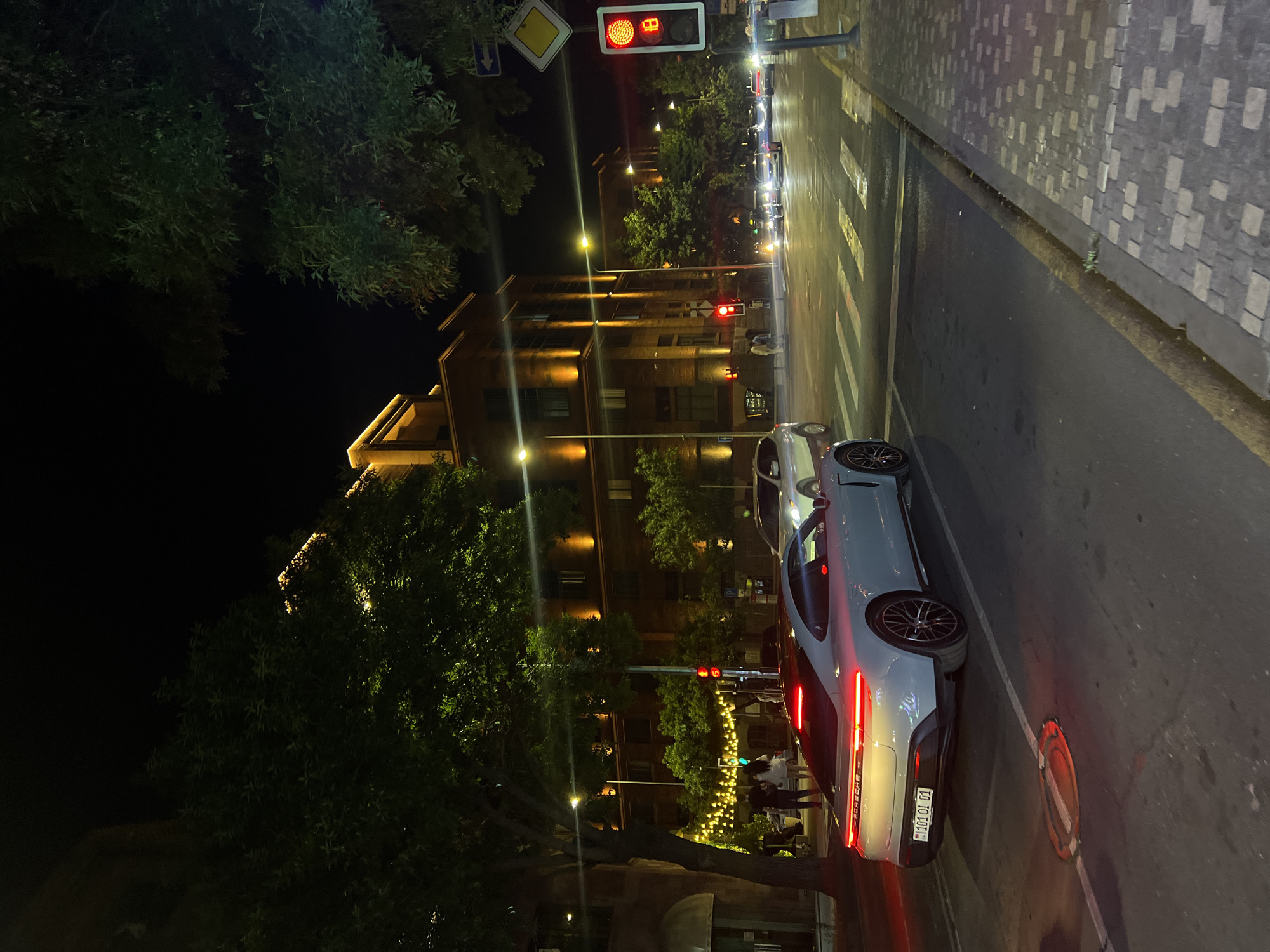 A street scene at night in Yerevan, Armenia, is depicted. A white Porsche Taycan with license plate "101 OL 01" is parked on the right side of the road. The car's brake lights are illuminated red. Across the street, a building with warm yellow lighting is visible, with trees and greenery in front. A traffic light shows red. Further down the street, other cars are present, and a pedestrian crossing is marked on the asphalt. The sidewalk is paved with stone.