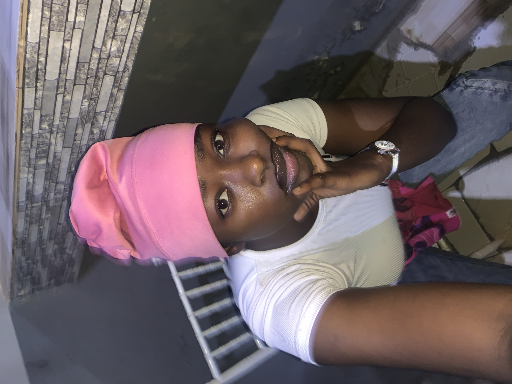 A person with dark skin, wearing a pink head covering and a white ribbed t-shirt, is photographed from a high angle. They are wearing a silver-colored wristwatch on their left wrist and have their fingers near their mouth. The background includes a wall with light gray rectangular tiles, a metal rack or frame painted white, and denim-colored clothing. Based on the location information provided, the image was taken in New Stadium, Nigeria.