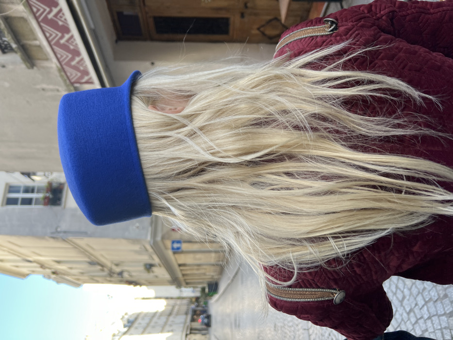 A person is depicted from a rear perspective, featuring long, wavy, blonde hair extending from beneath a bright royal blue, felt-like, pillbox-style hat. The individual wears a maroon-colored, quilted jacket with distinctive light-brown, woven-patterned shoulder straps adorned with small metallic buttons or studs.
The scene takes place on a cobblestone street in the Le Marais district of Paris, France. In the background to the left, a narrow street recedes, bordered by multi-story, light-colored stone buildings typical of Parisian architecture, featuring multiple windows. A blue square street sign with a white symbol is affixed to a building wall. A dark-colored vehicle is visible further down the street. To the right, an older building with a prominent wooden double door is present. Above this door, a distinctive reddish-brown and white geometric patterned frieze is visible. The environment is brightly lit by natural sunlight, indicating a clear day.