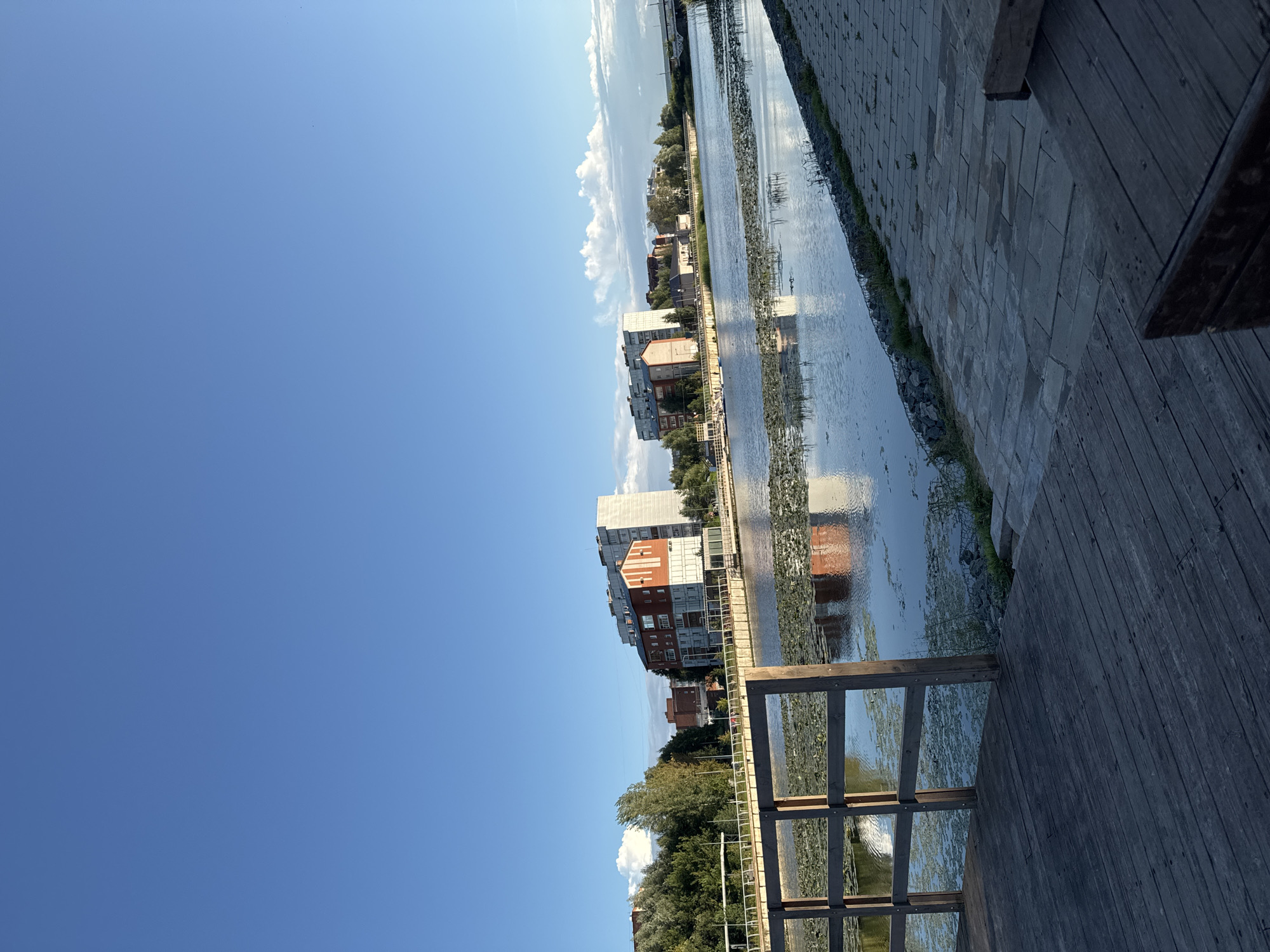 The image depicts a serene waterside scene in Votkinsk, Russia, likely during the late afternoon or early evening. In the foreground, a wooden deck or pier leads towards the water, its weathered planks forming a textured surface. To the left, a wooden railing adds a touch of rustic charm.
The water body in the midground is calm, reflecting the sky and the buildings on the opposite bank. Lily pads and aquatic vegetation dot the surface, creating a natural, somewhat unkempt appearance. Across the water, a series of modern buildings line the embankment. Notable among them is a multi-story building with striking red and white vertical stripes, suggesting a contemporary architectural style. Other buildings, varying in height and design, are also visible, interspersed with trees. A bridge can be seen in the distance, spanning the waterway.
The sky above is a vibrant, clear blue, with a few wispy white clouds adding depth and interest. The overall atmosphere is peaceful and tranquil, capturing a quiet moment in an urbanized natural setting. There are no people or discernible actions in the image, focusing the viewer's attention on the interplay of architecture, water, and sky.