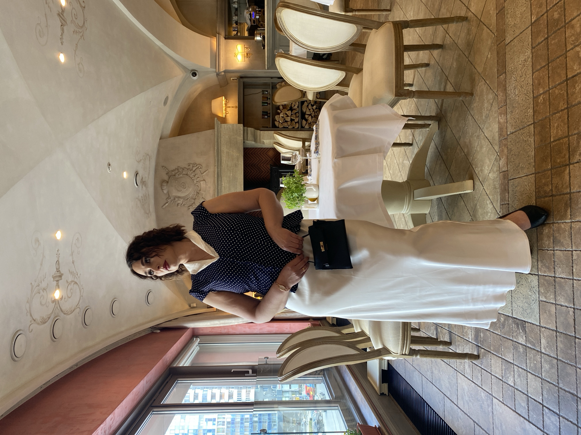 A woman stands inside a room with an arched ceiling and elaborate wall decorations. She is wearing a navy blue polka dot top and a long white skirt, with a black purse at her side. The room appears to be a restaurant, indicated by the set tables with white tablecloths and chairs. Outside the window, a cityscape is visible, suggesting an urban location. The floor is tiled in a patterned design.