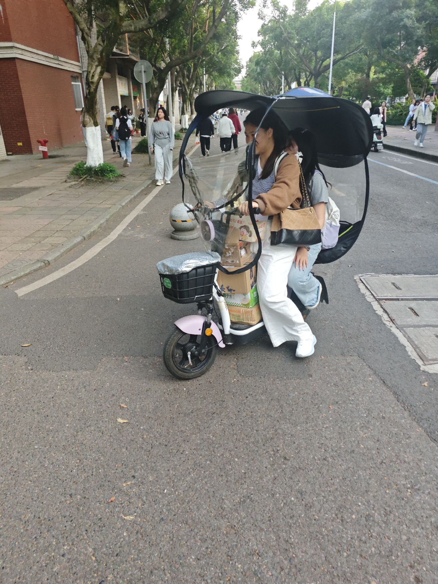 The image shows two people riding a pink electric scooter with a black canopy on a paved road. They are likely in the city of Chongqing High-tech Zone, China, based on context. The rider is wearing a brown jacket, striped shirt, and white pants. The passenger wears a gray shirt and light blue jeans. They are both carrying bags. The scooter has boxes and a covered basket in front. Pedestrians walk on the sidewalk beside a building with red brick in the background. Trees line the street.