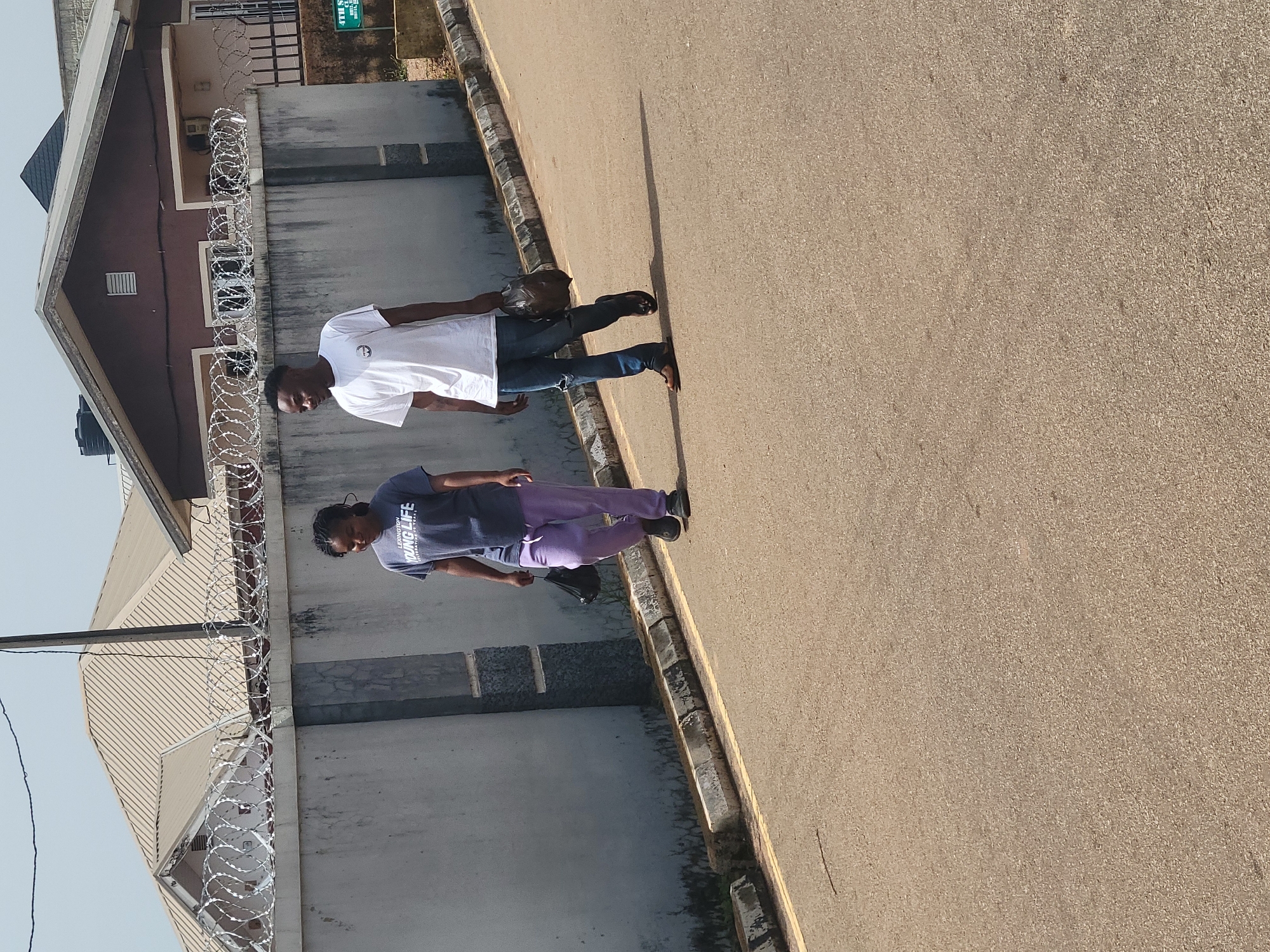 In the city of Jos, Nigeria, two individuals walk side-by-side on an asphalt road. The person on the left, a woman wearing a dark blue t-shirt with white lettering and light purple sweatpants, carries a black plastic bag in her right hand. The person on the right, a man, is dressed in a white t-shirt and blue jeans with a rip on the left knee, and also carries a dark plastic bag in his right hand. They are walking away from the viewer towards a grey concrete wall topped with coiled barbed wire, which separates the road from a residential area. Beyond the wall, two houses are visible, one with a light brown, angular roof and the other with a darker brown, gabled roof. The sky is clear and bright, suggesting a daytime scene. A signpost with green text is partially visible on the right side of the image.